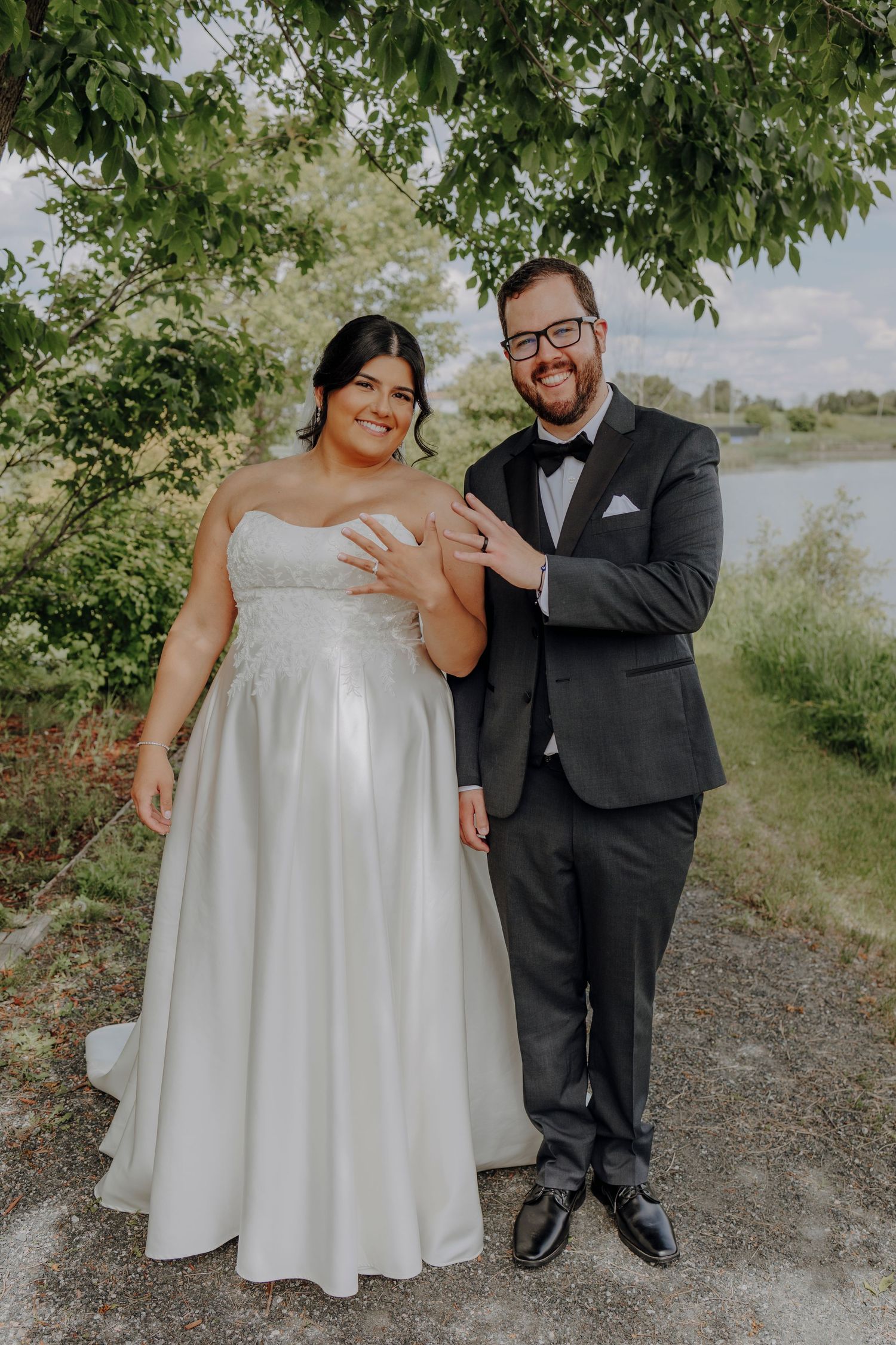 A couple in wedding attire poses together outdoors on a gravel path surrounded by green trees on a sunny day.