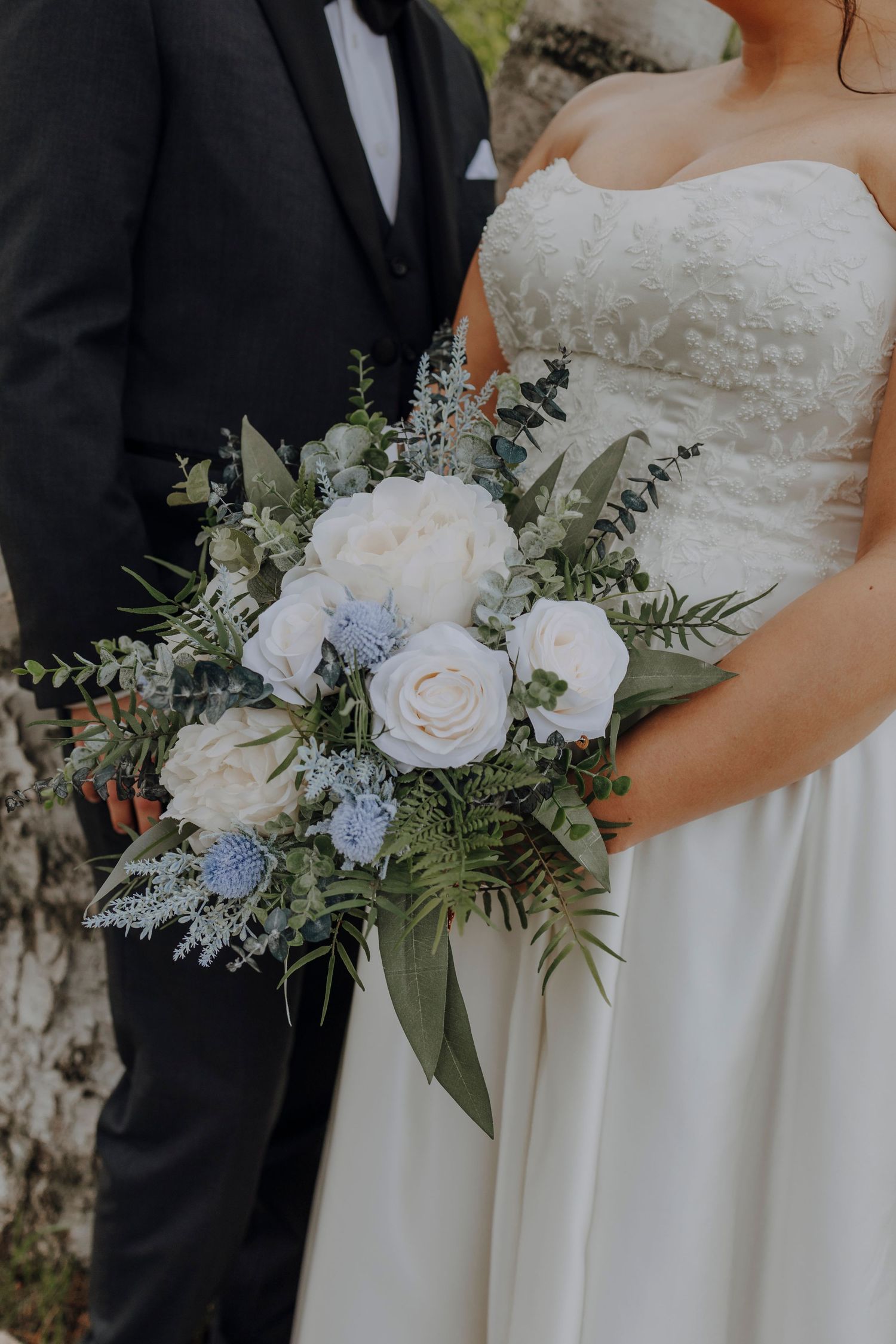 A wedding bouquet with white roses, blue flowers, and greenery held against a white dress and black tuxedo.