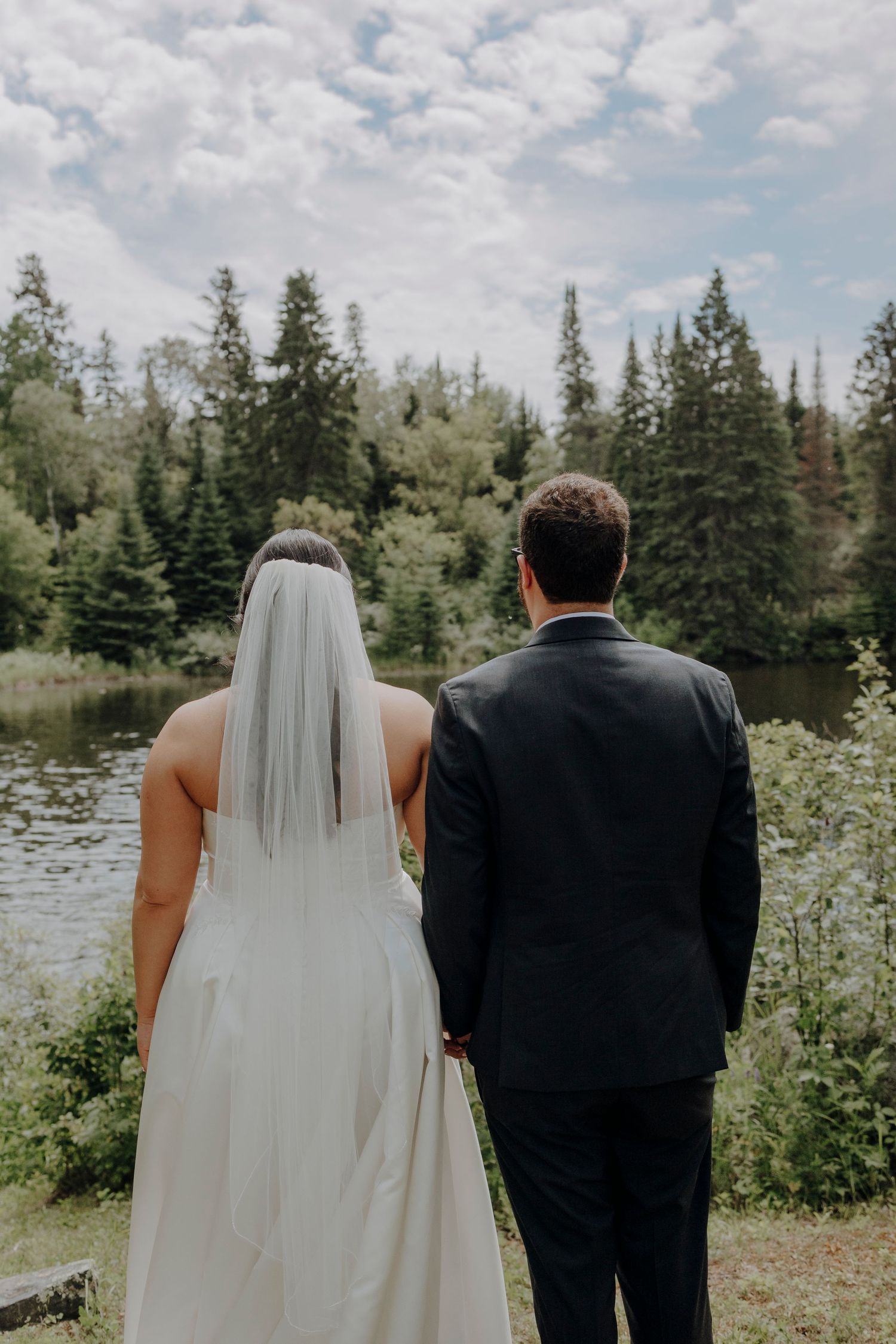 Back view of couple in wedding attire looking out at scenic mountain landscape with evergreen trees.