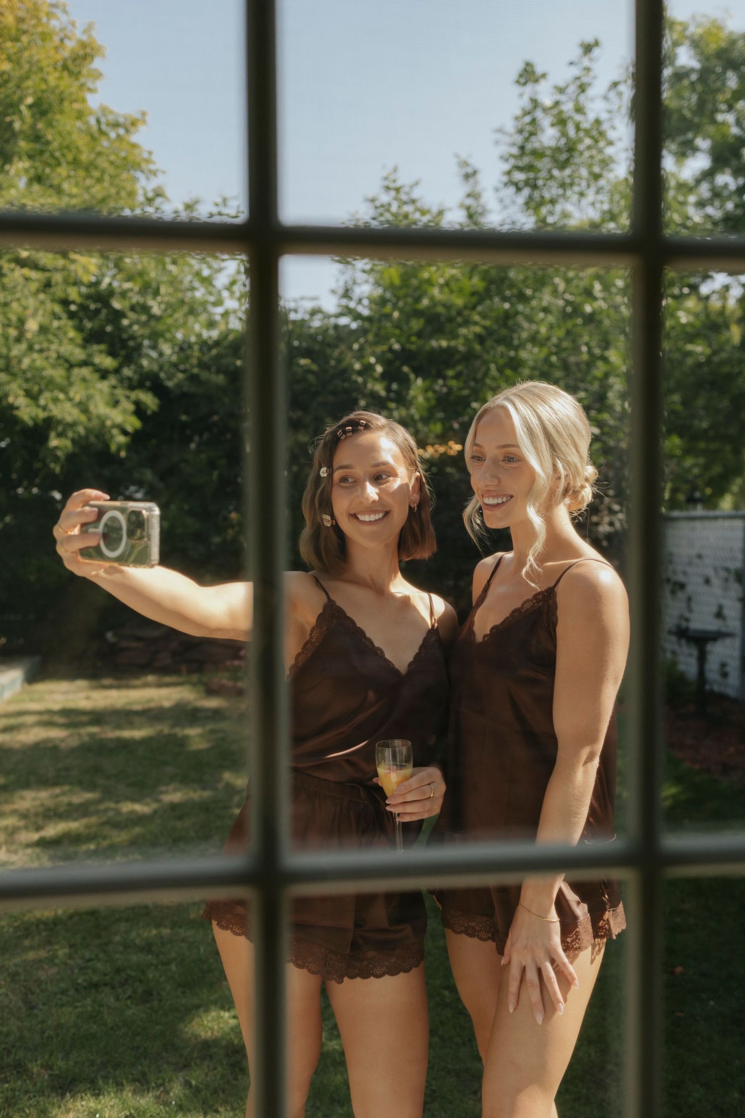 Two friends in brown dresses laughing and taking photos together in a sunny outdoor setting.