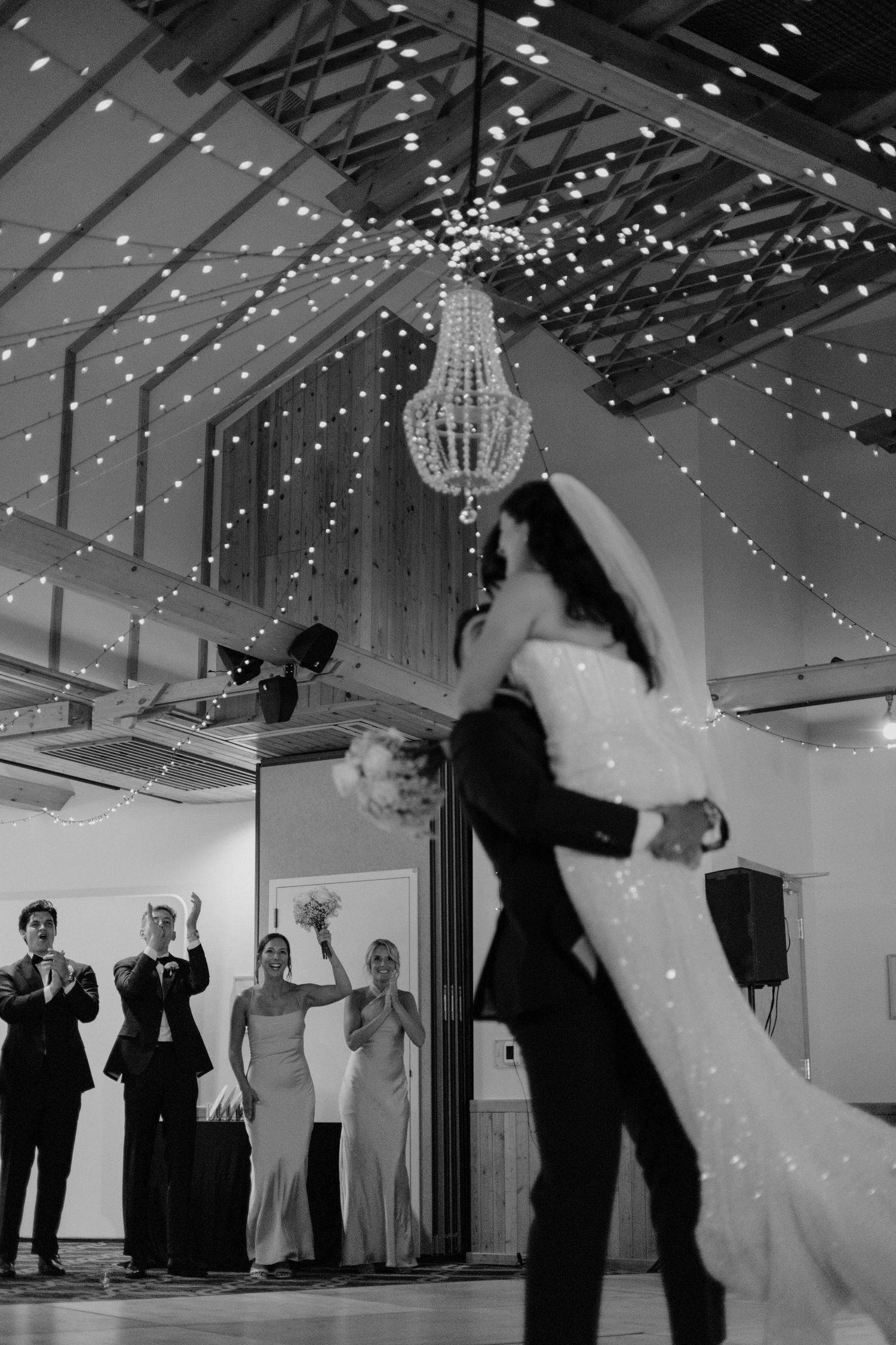 Wedding guests dance and celebrate under twinkling string lights at an indoor reception venue.