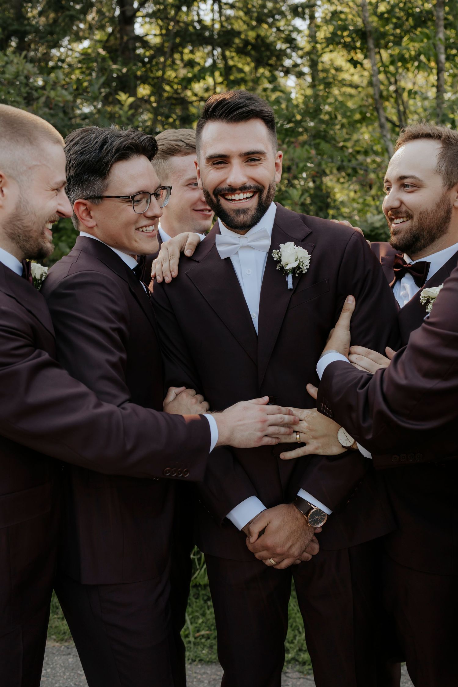 Groomsmen in burgundy suits sharing a candid moment and laughing together outdoors.