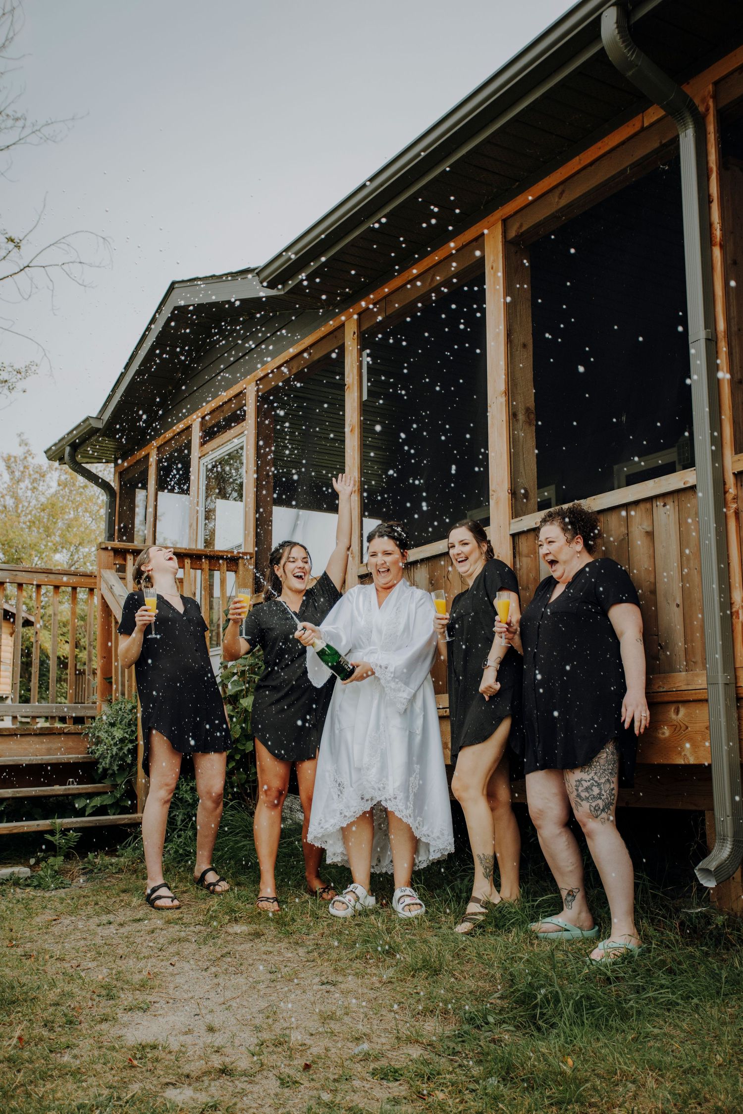 Wedding party celebrating with champagne spray outside rustic wooden cabins on a grassy lawn.
