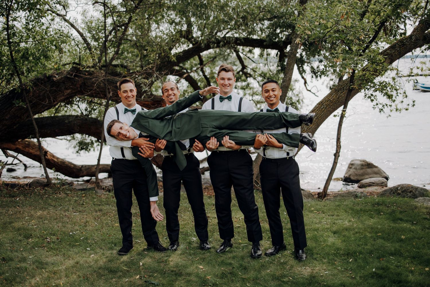 Groomsmen in suspenders and blue ties pose creatively under a large tree branch by the water.