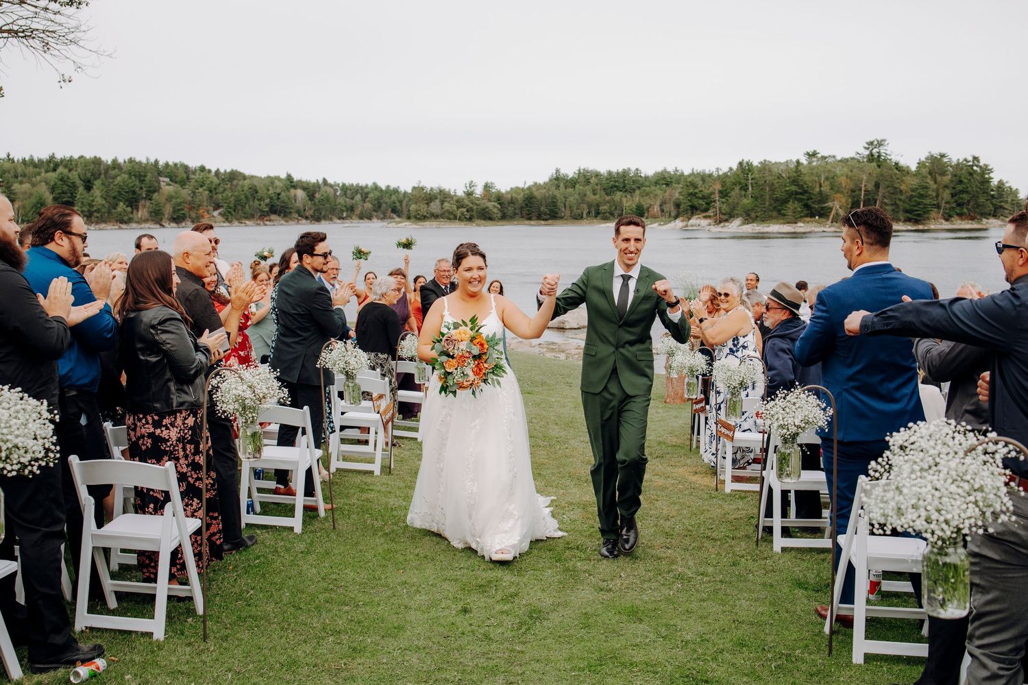 Outdoor lakeside wedding ceremony with white chairs and guests gathered under open sky.