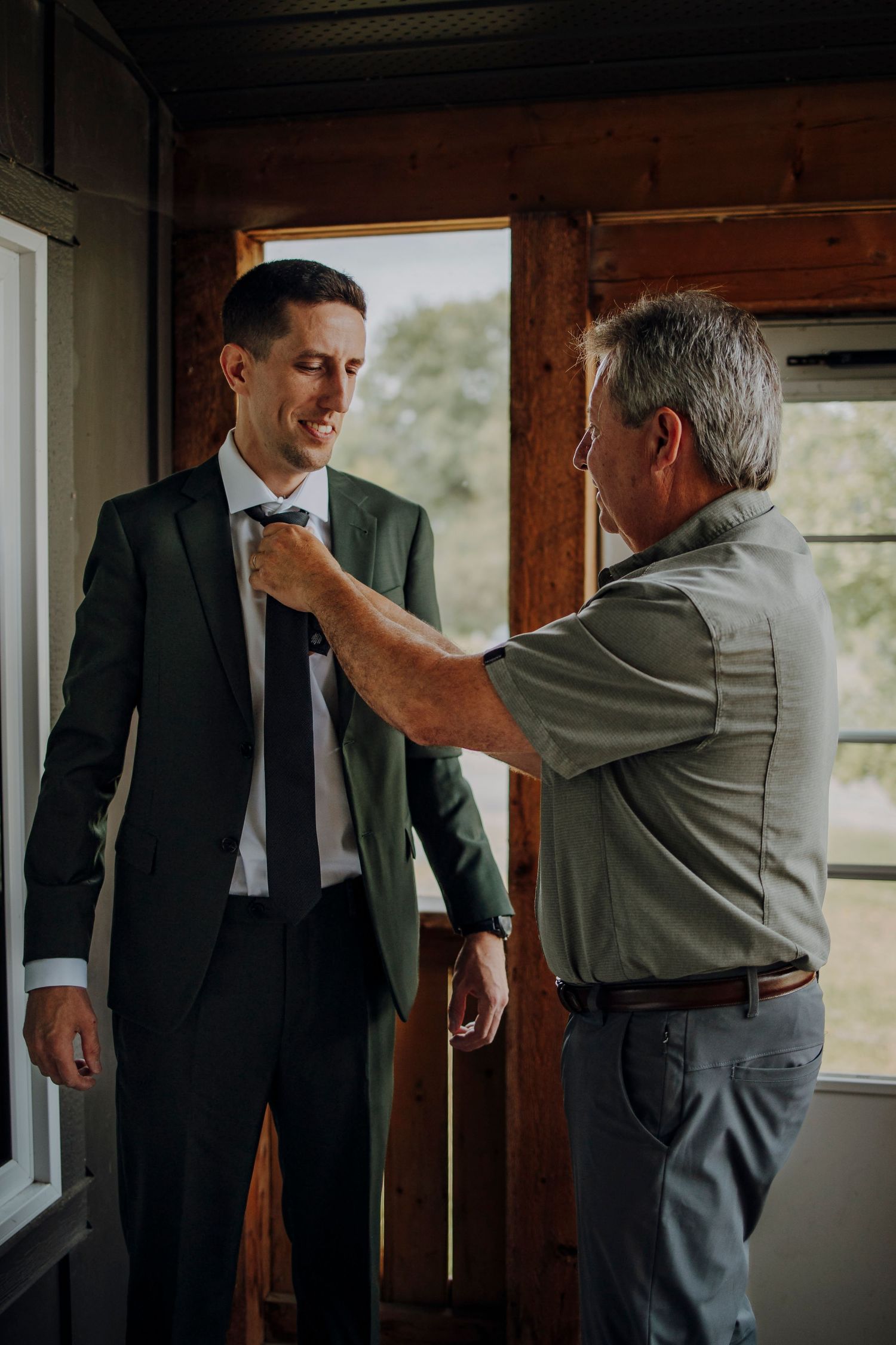 Two people in a rustic room preparing for a formal event with one helping adjust the other's attire.