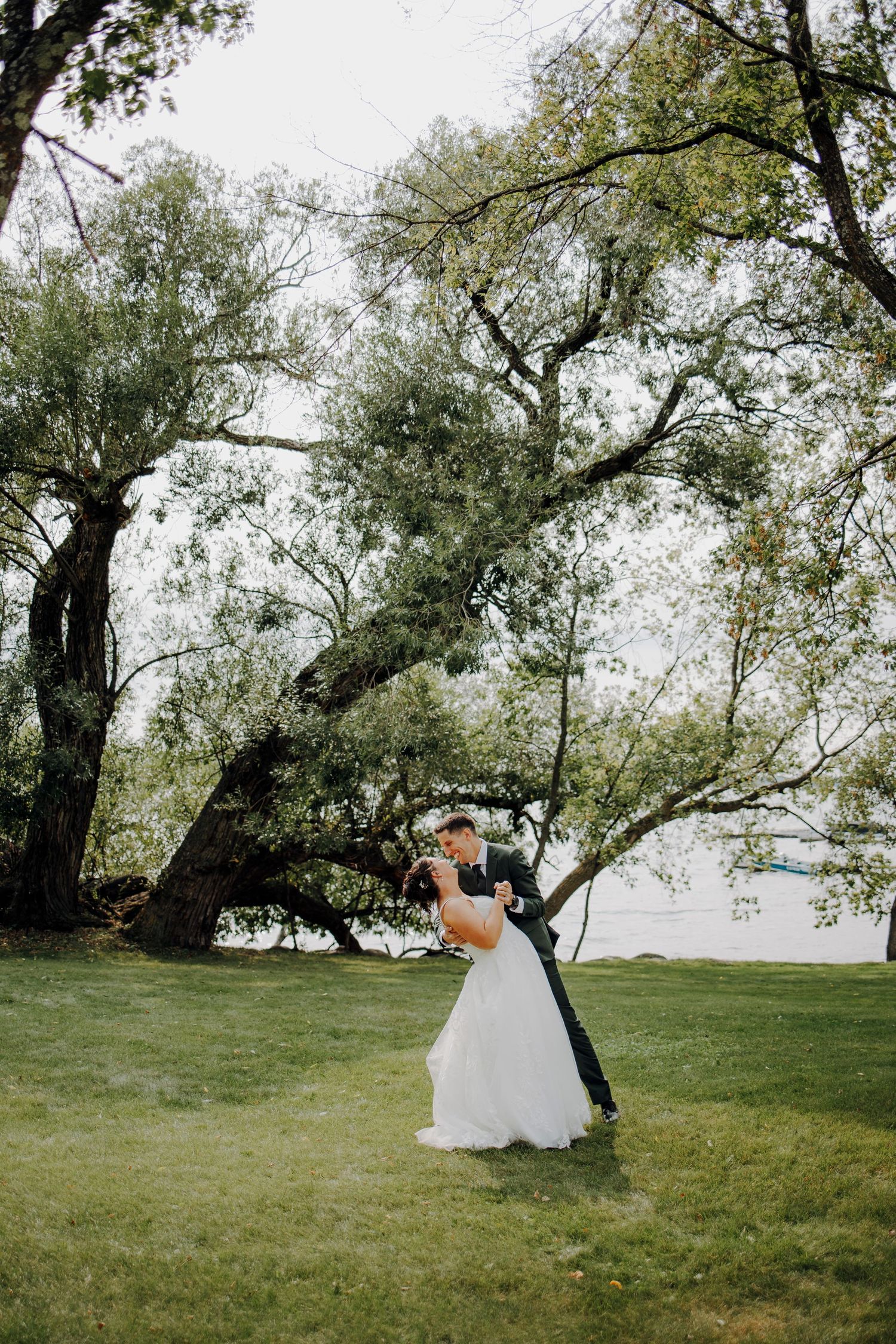 Wedding ceremony setup under tall trees by a lakeside with white chairs and natural scenery.