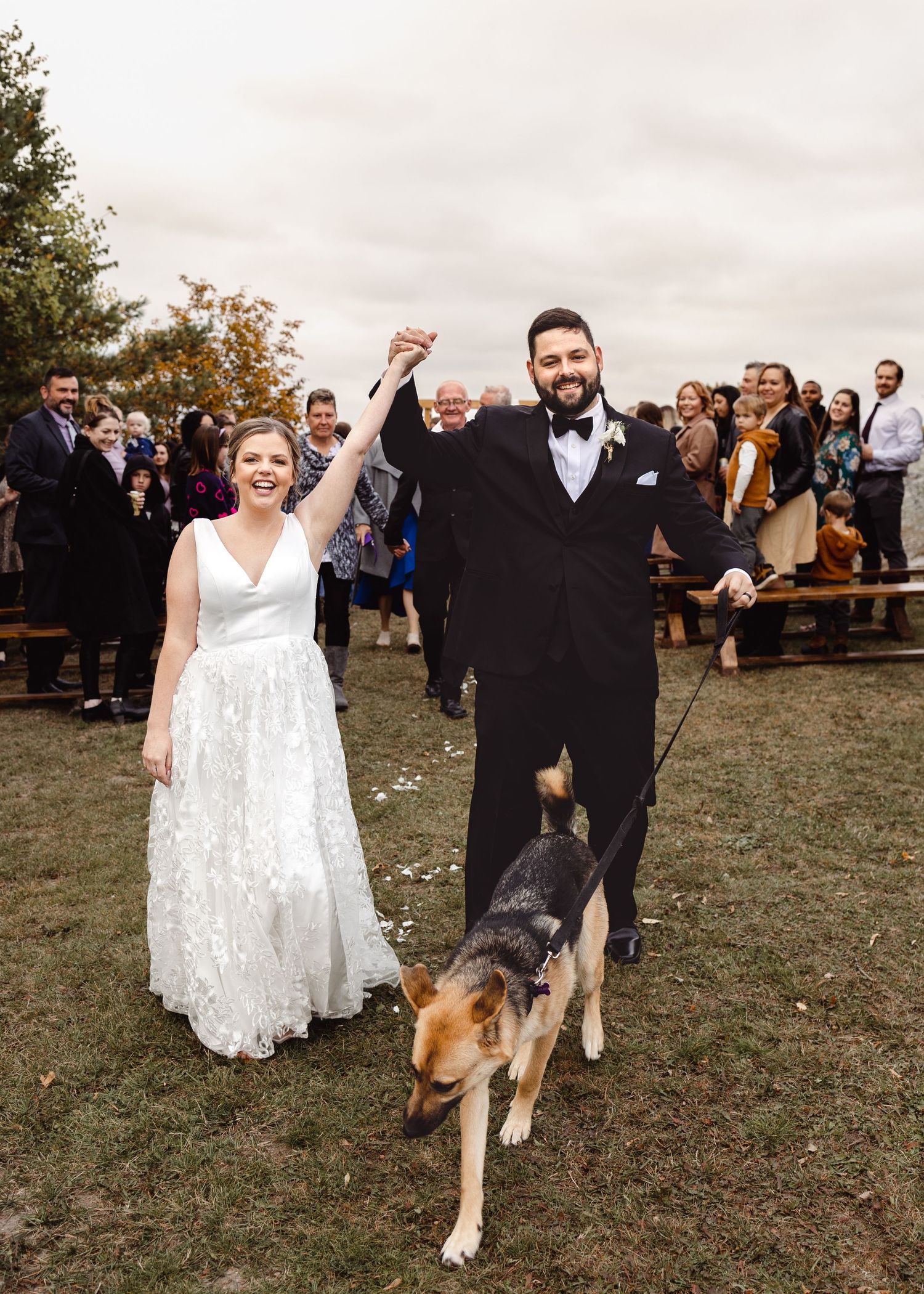 Newlyweds walk with their dog during their outdoor wedding celebration as guests cheer in the background.