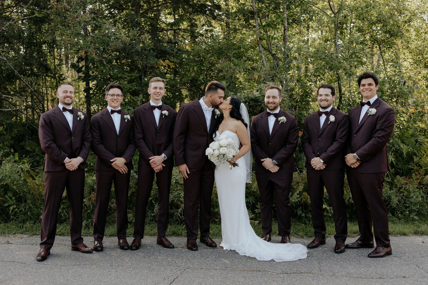 Wedding party in burgundy suits poses with bride outdoors.