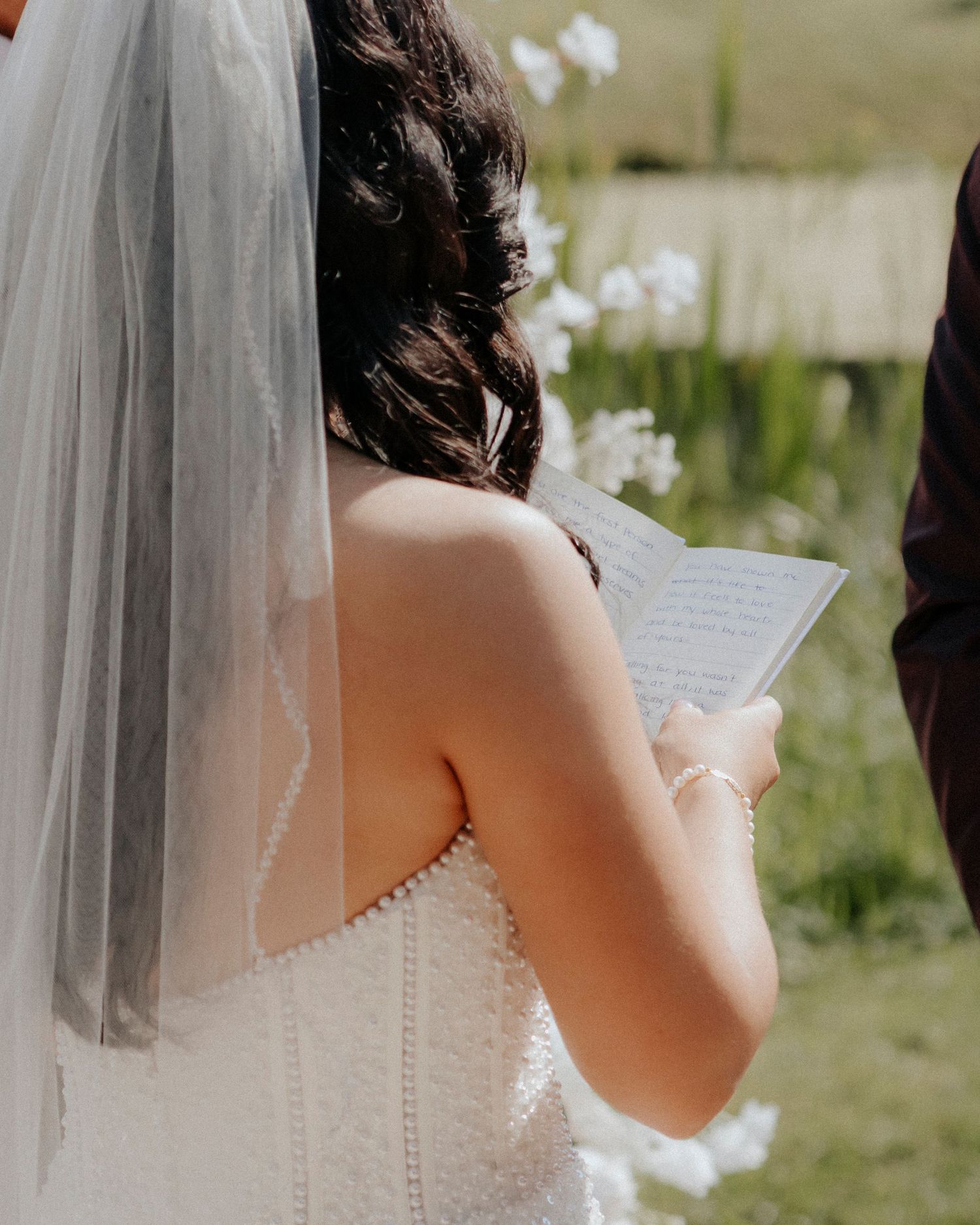 Close-up of a bride in a veil reading from paper during an outdoor ceremony.