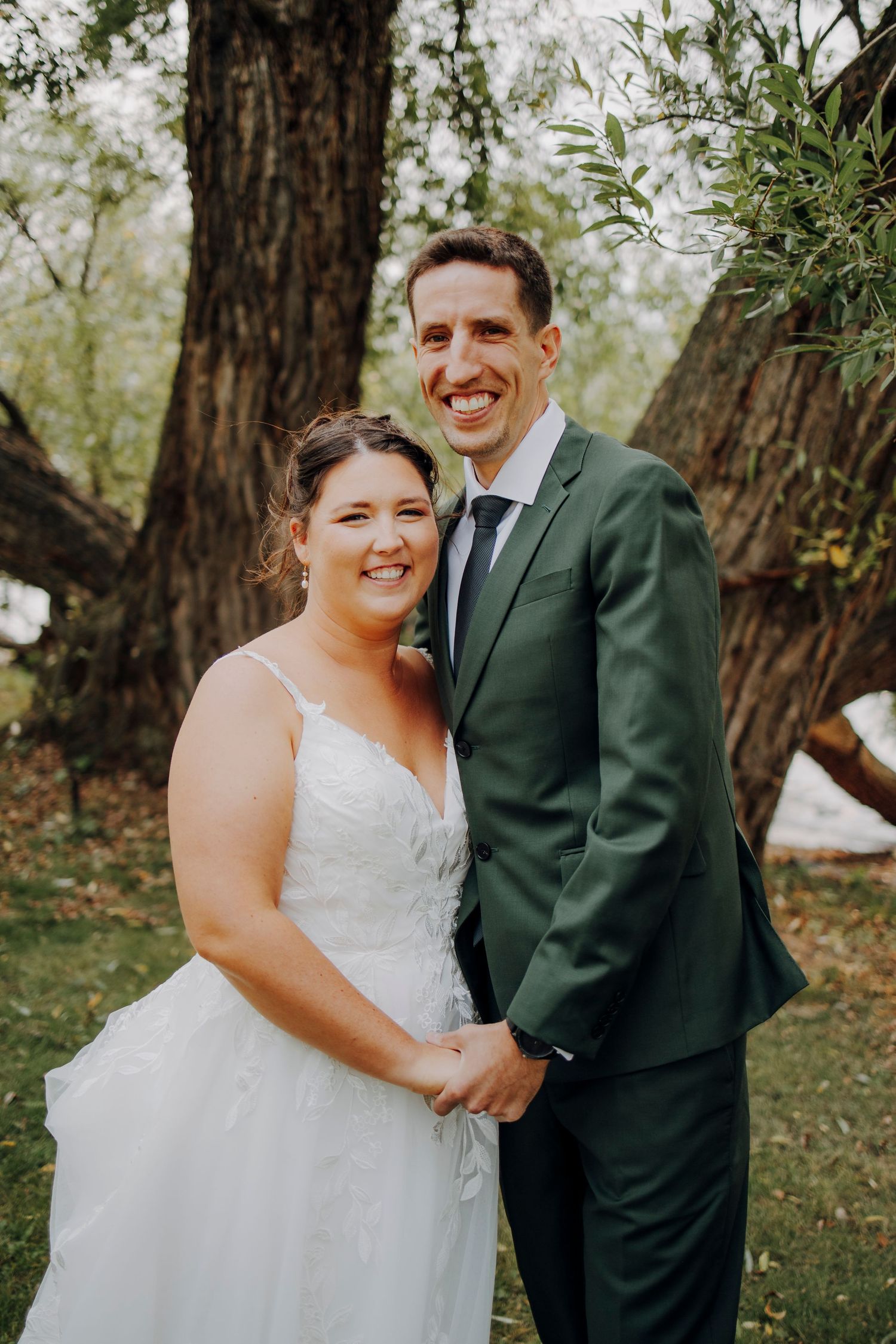 A joyful wedding couple embraces outdoors among trees wearing a white gown and dark green suit.