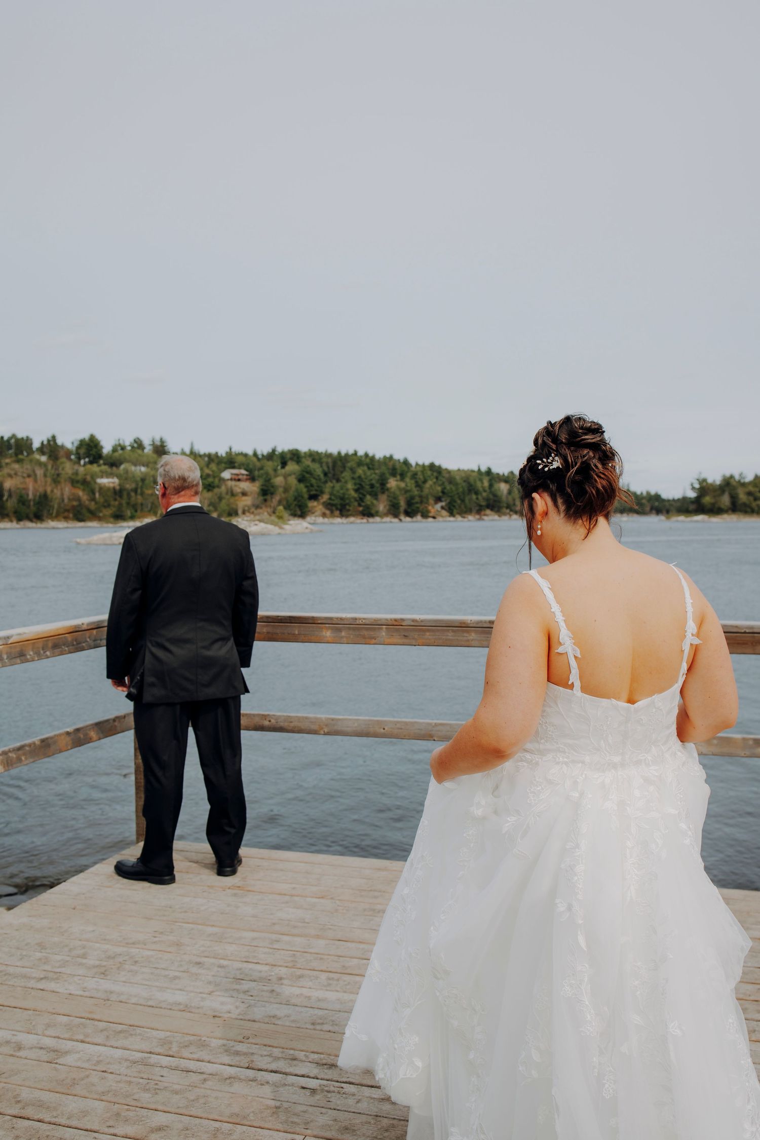 A couple in wedding attire walks along a wooden dock overlooking a serene lake on their special day.