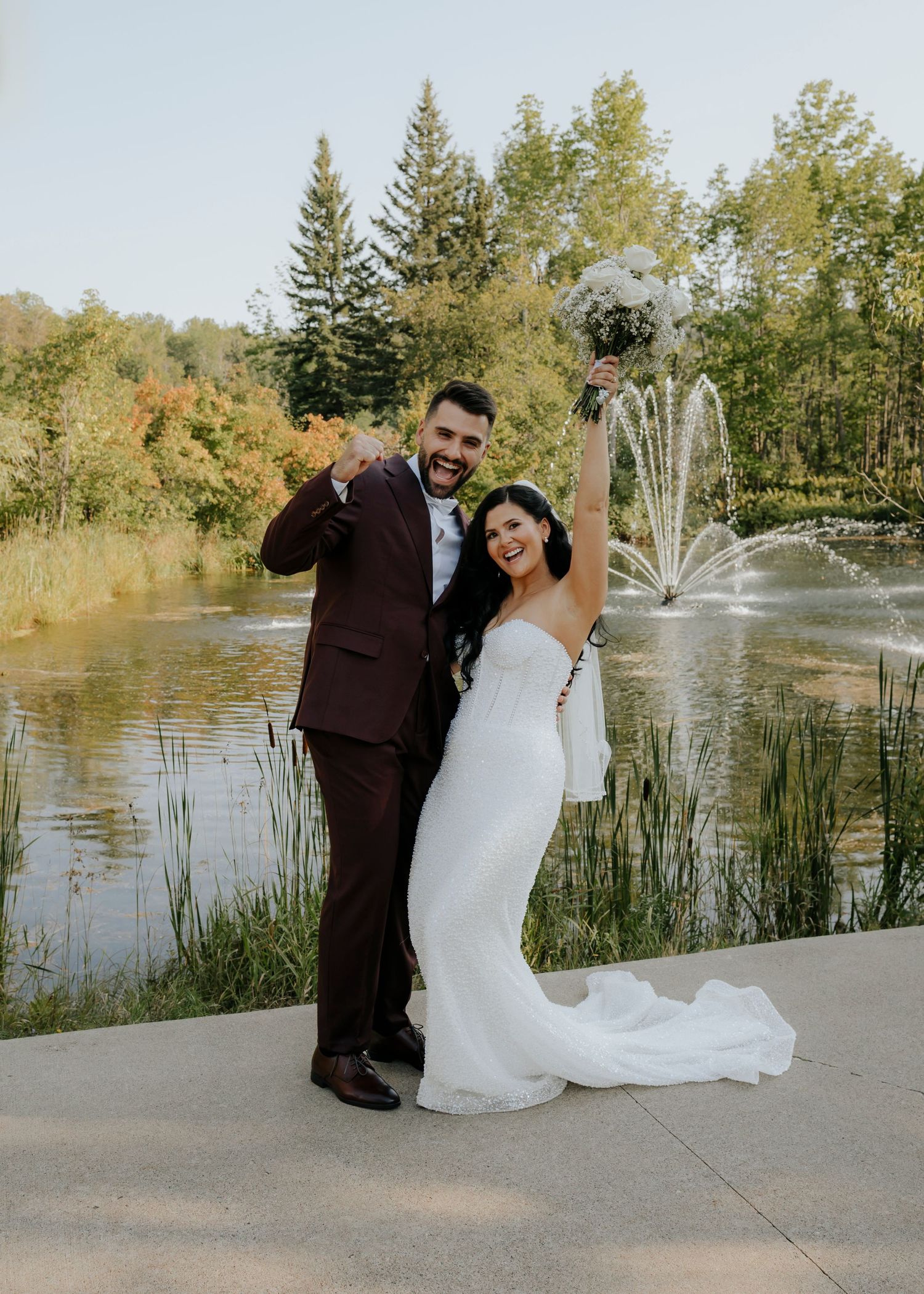 Wedding couple poses by lakeside fountain in formal attire.
