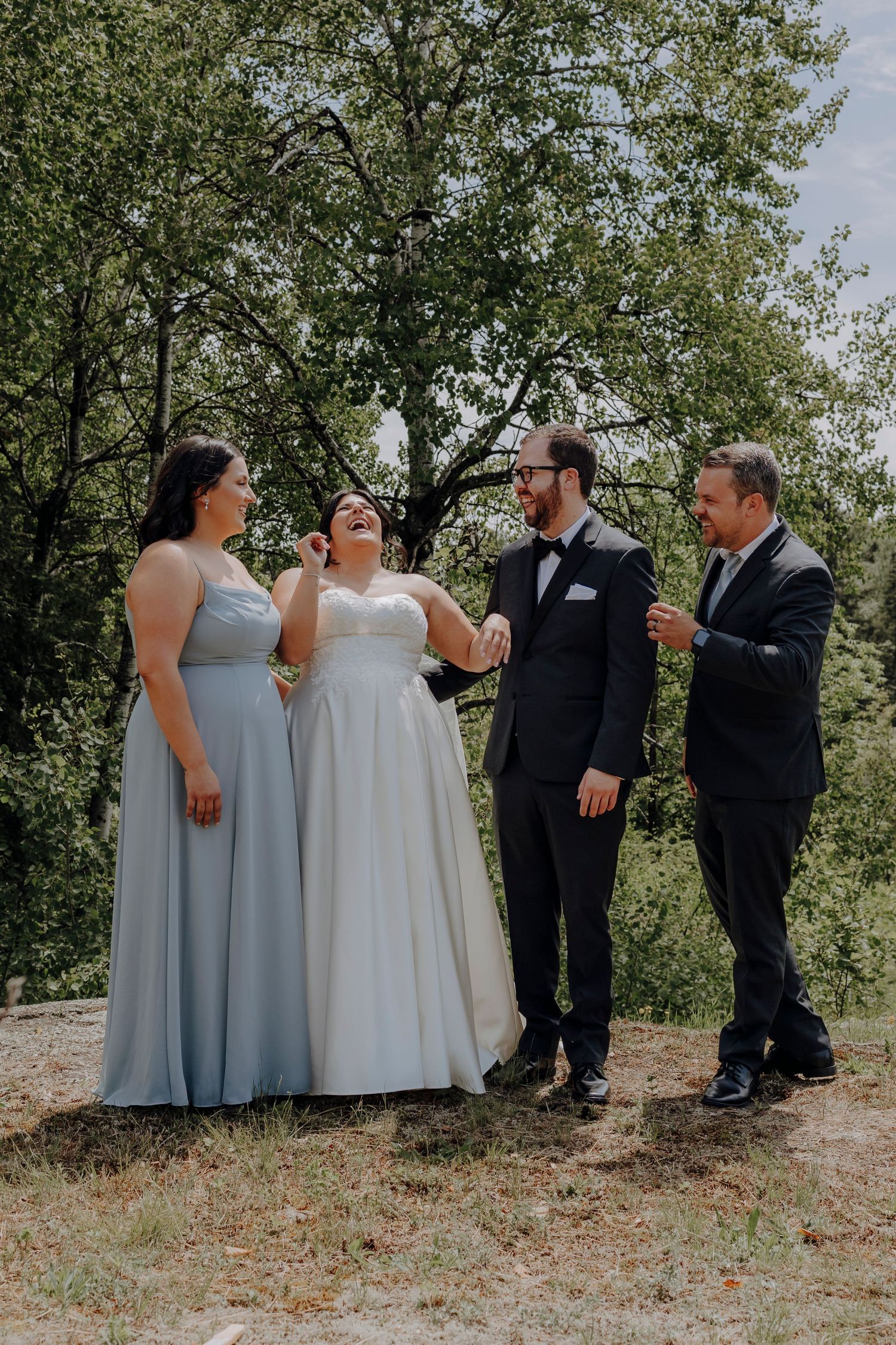 Wedding party in formal attire poses playfully outdoors under green trees on a sunny day.