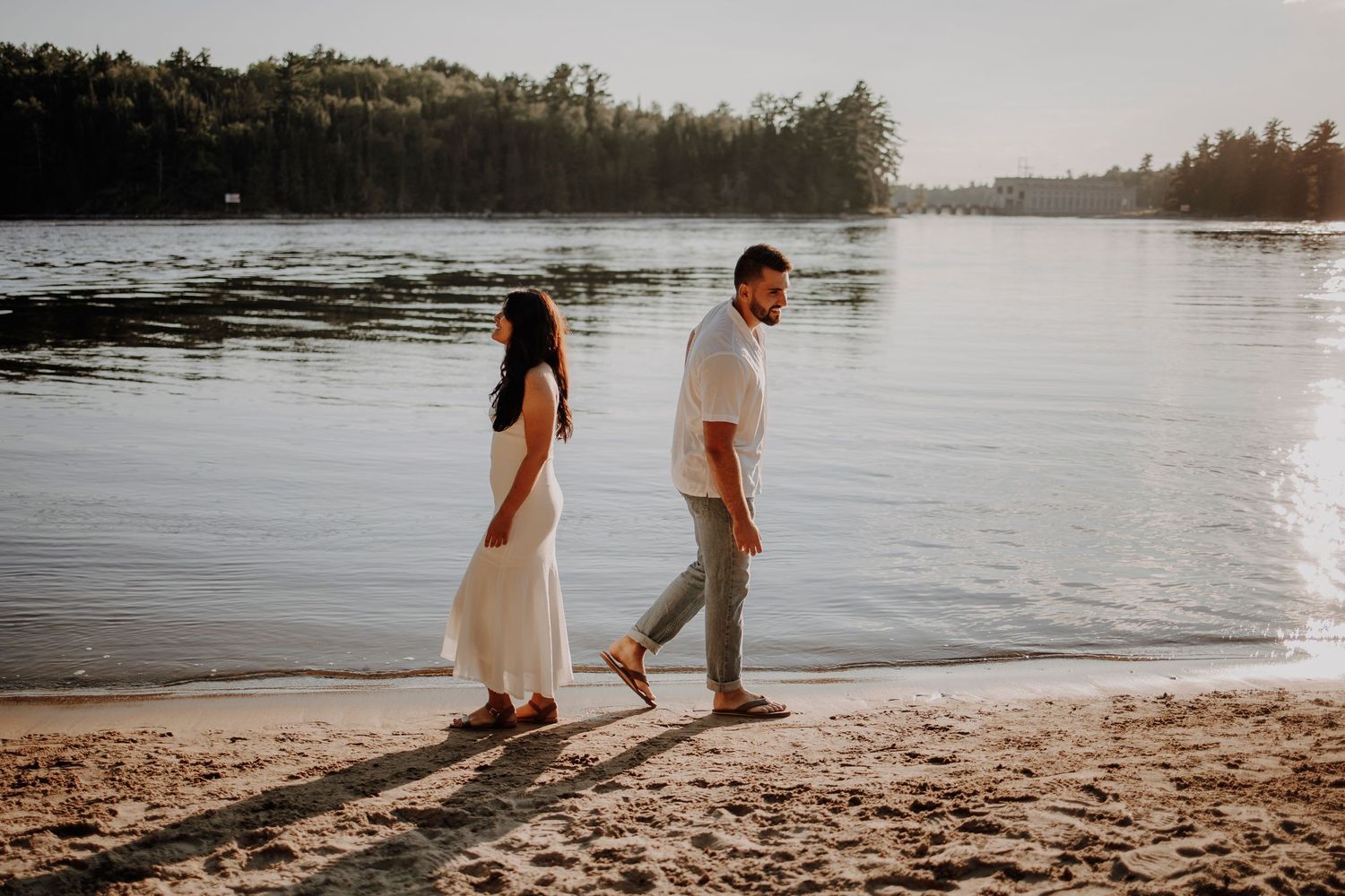 A couple walking along a peaceful lakeshore beach at sunset.