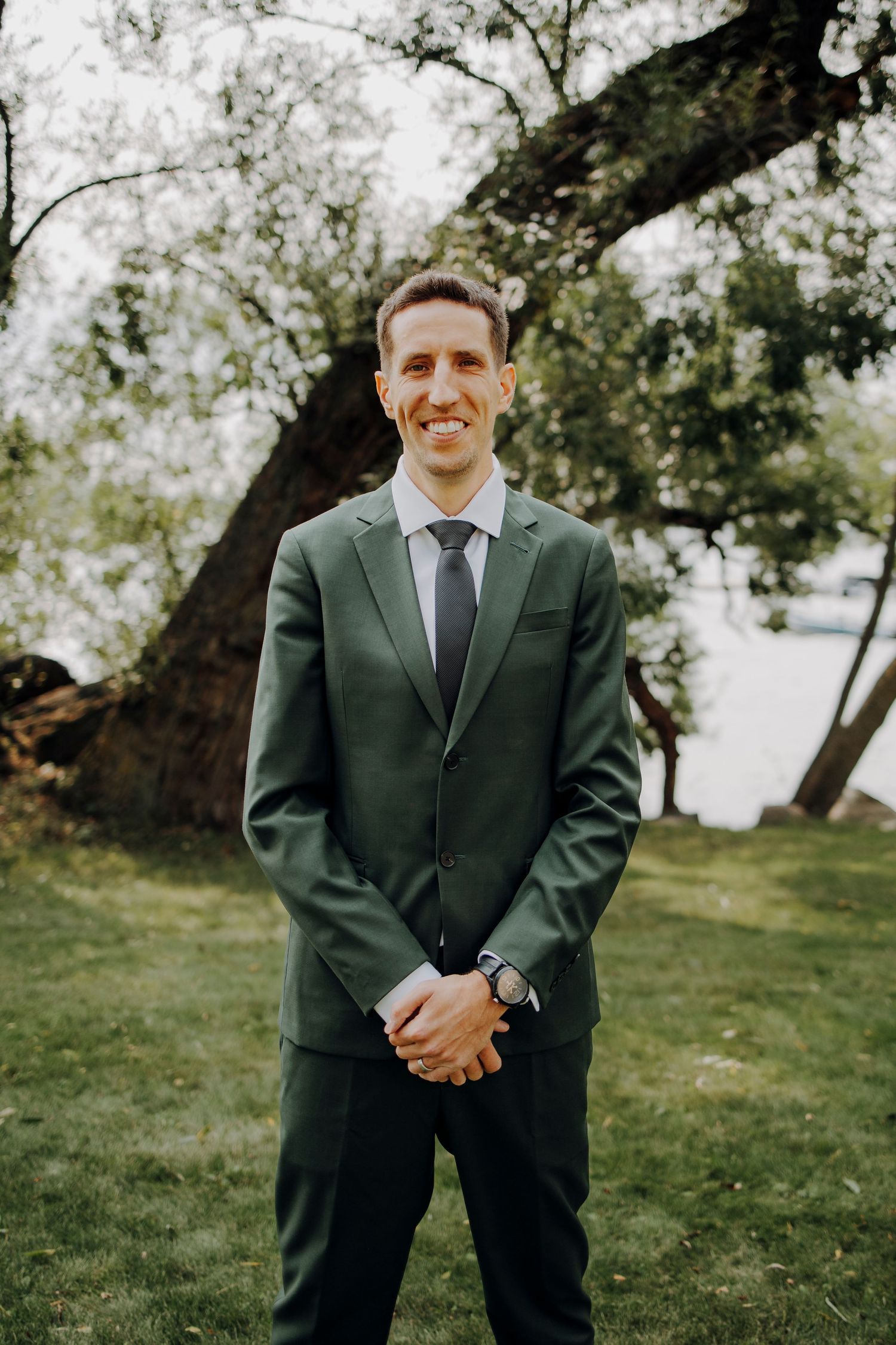 Groom in dark green suit smiles while standing in outdoor garden setting.