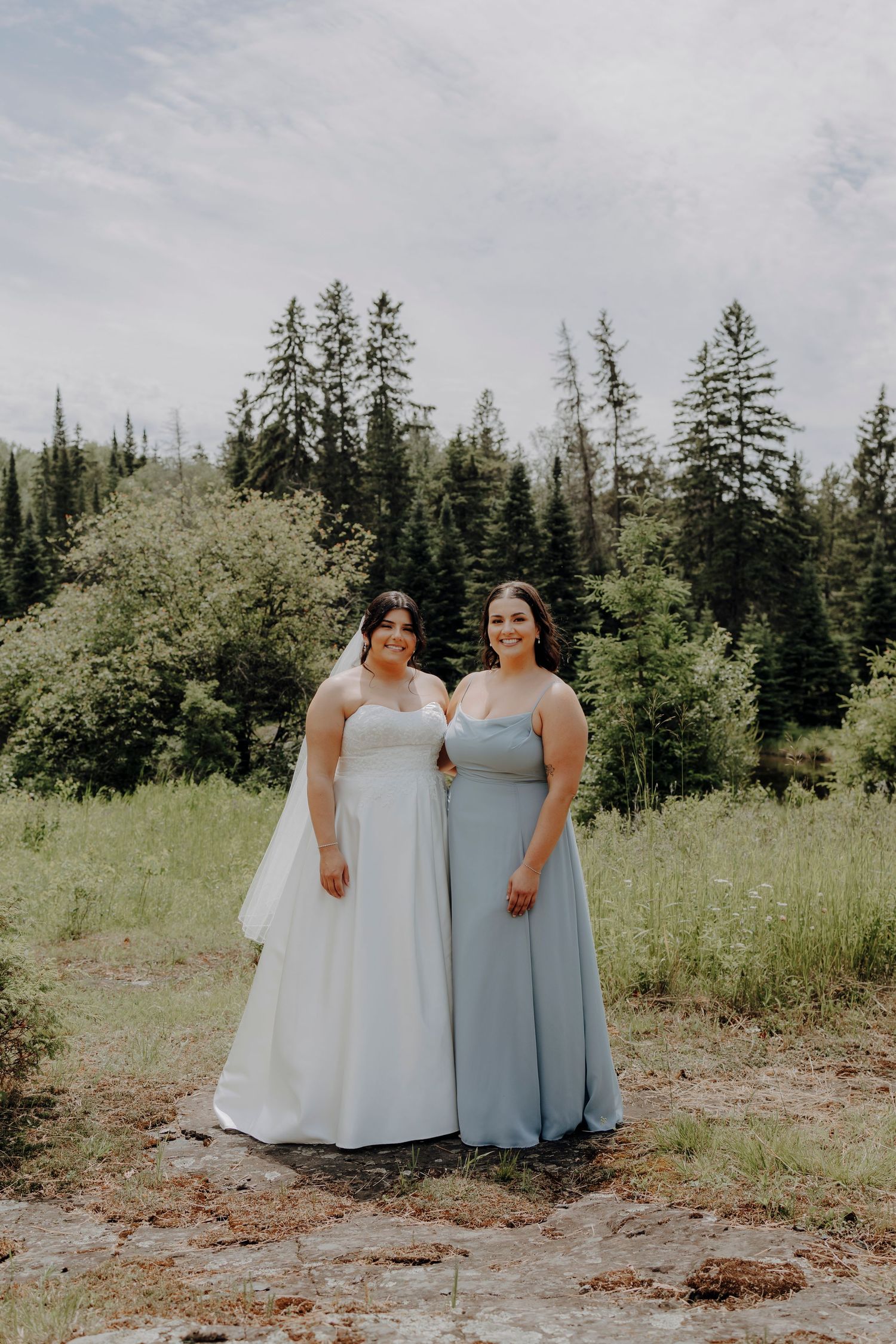Two people in coordinating light blue and white formal dresses stand together in a scenic outdoor setting.