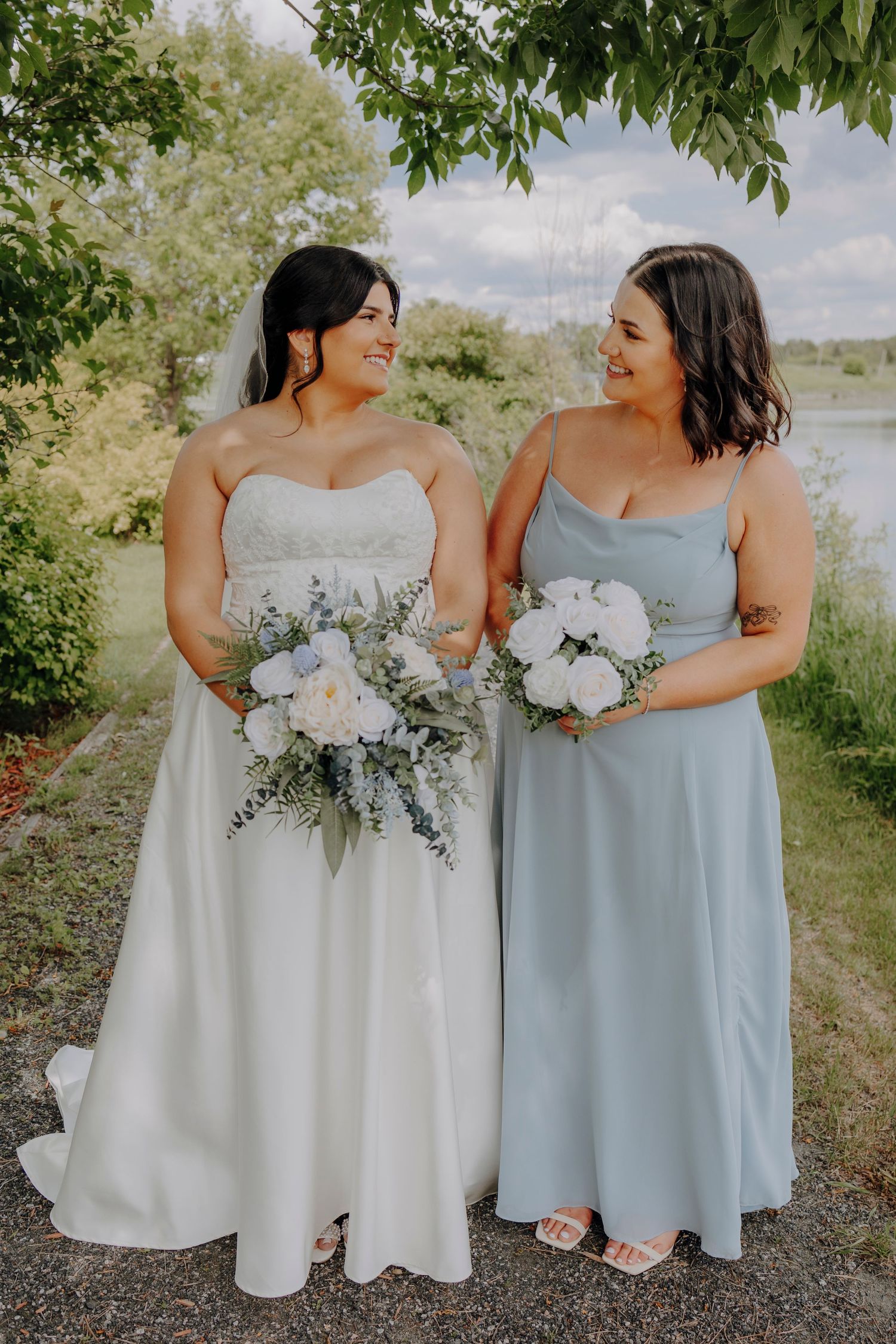 Bride in white gown and bridesmaid in light blue dress stand together holding matching floral bouquets.