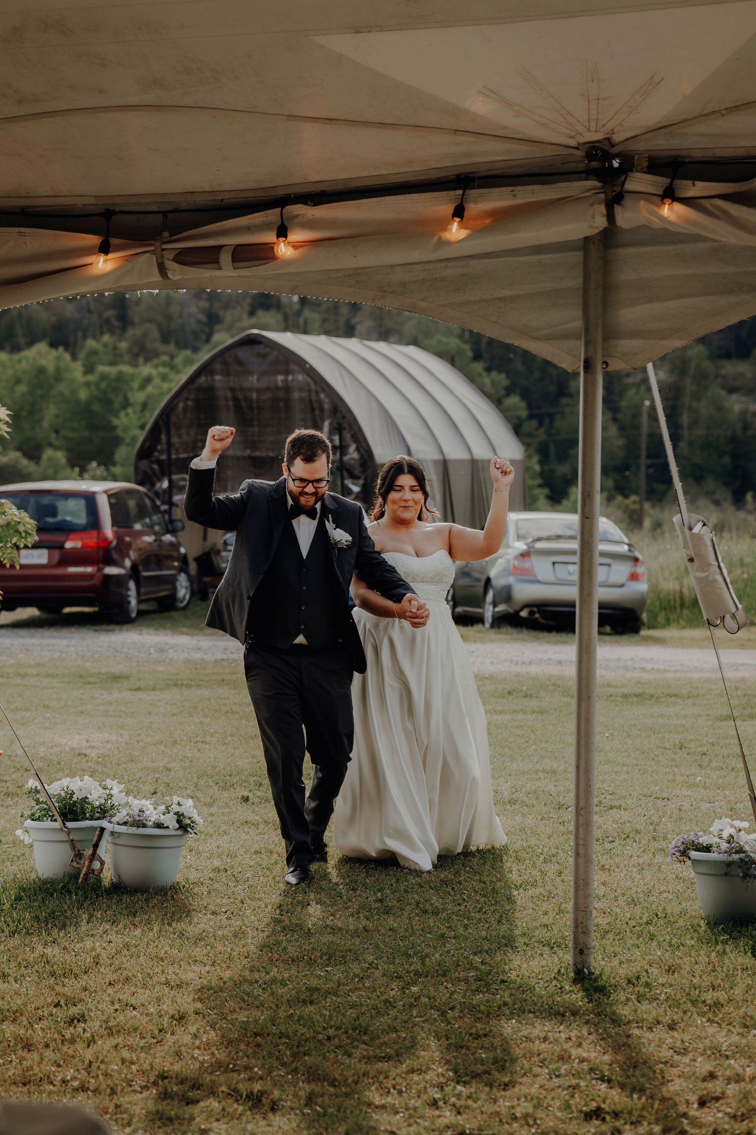 A couple celebrates under a tent canopy at a rustic outdoor wedding with string lights and potted flowers.