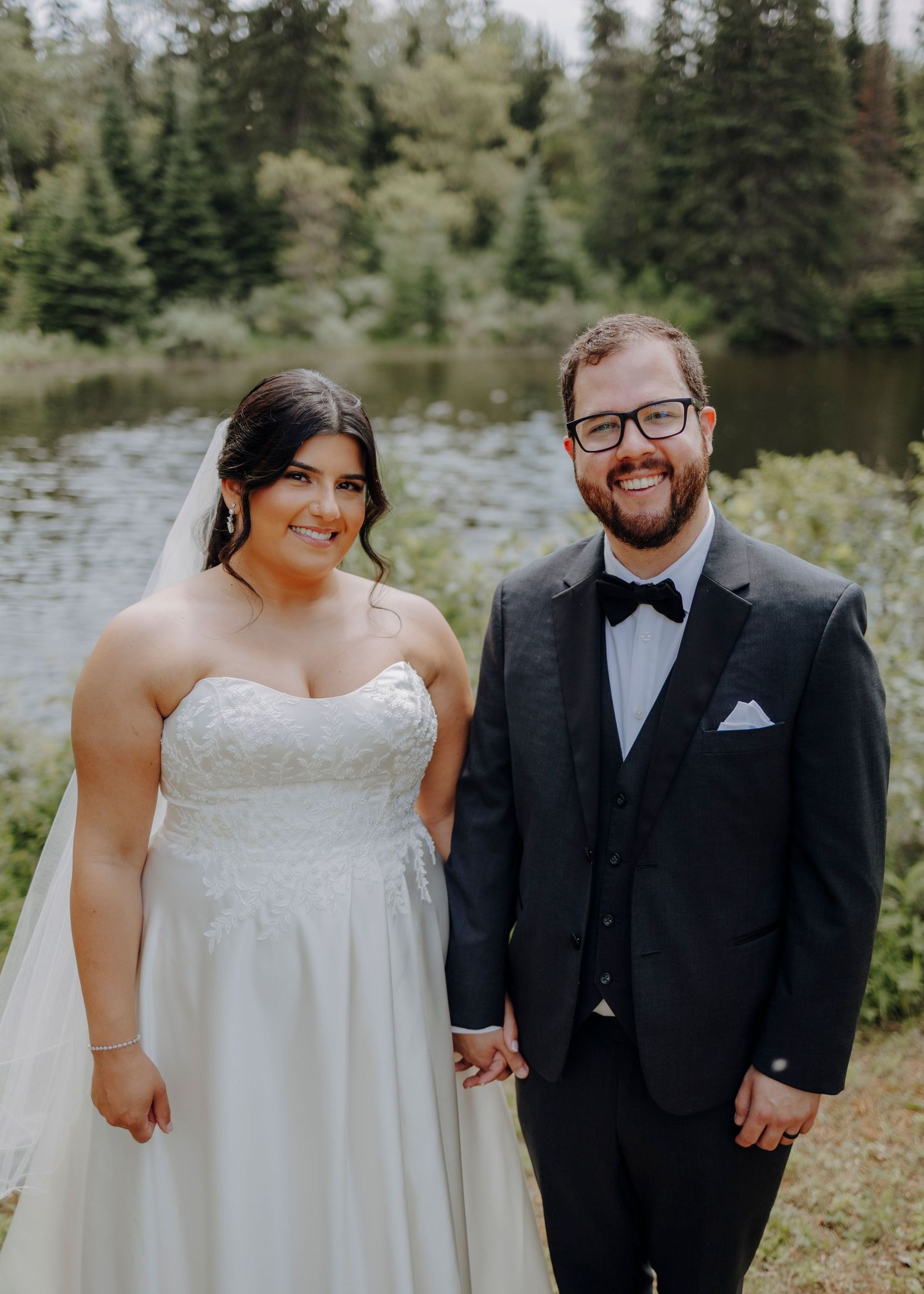 A wedding couple poses together outdoors with evergreen trees and a lake in the background.