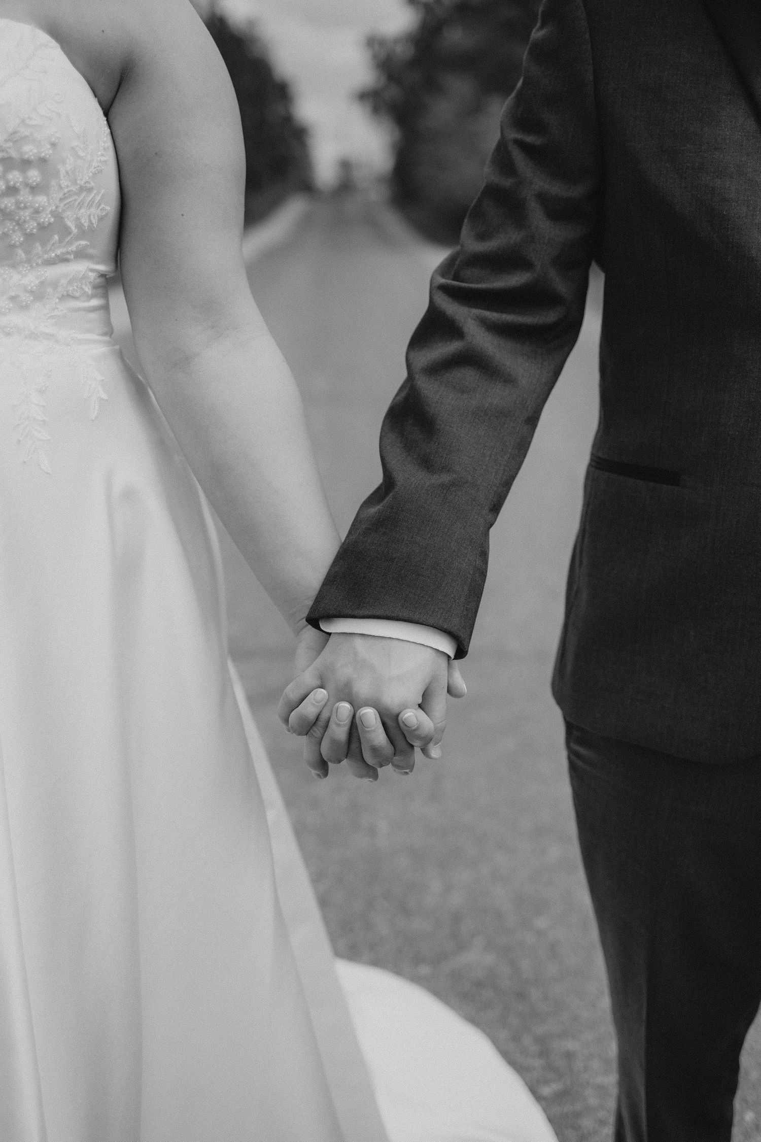 Close-up black and white photo of a bride and groom holding hands on their wedding day.