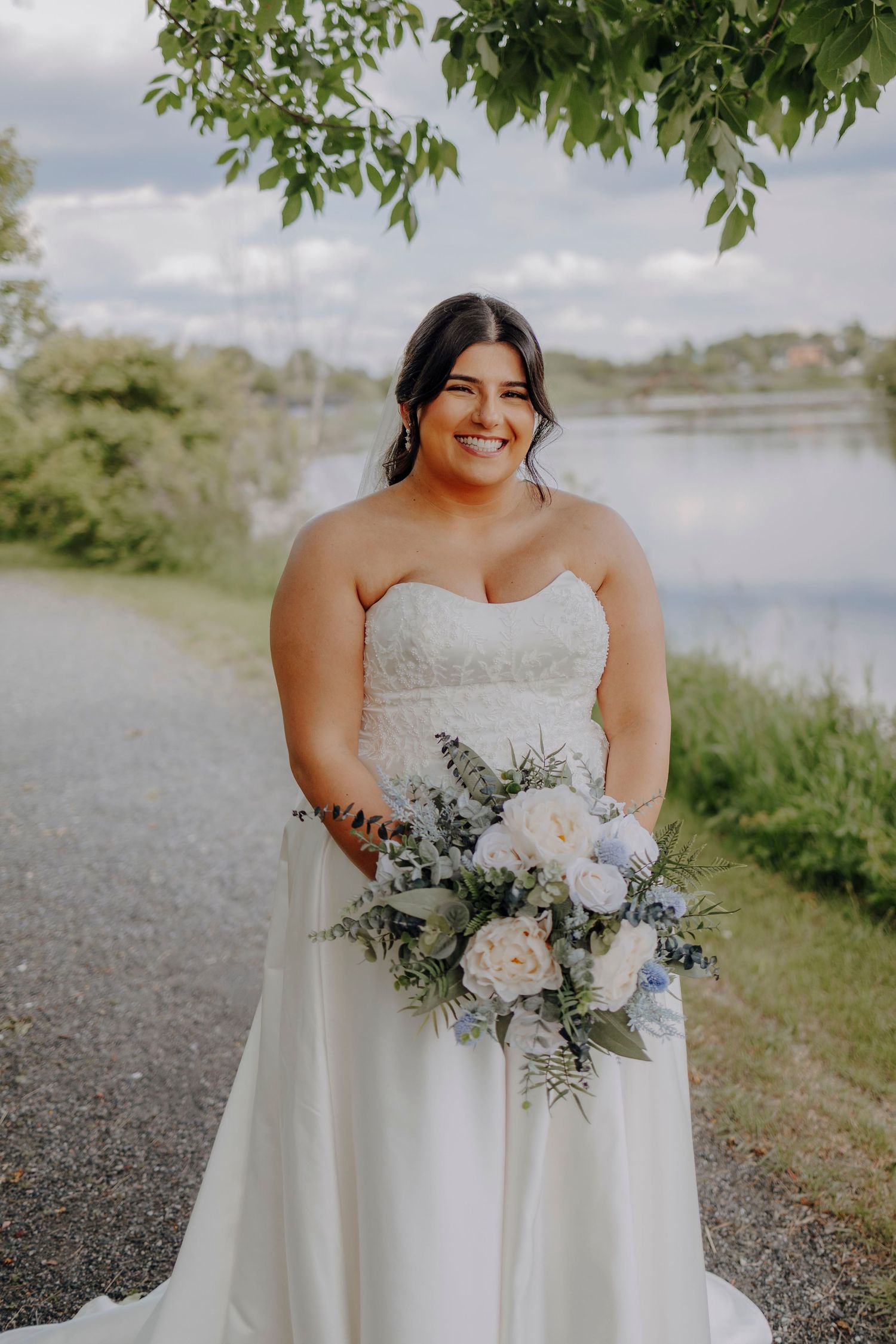A bride in a strapless white wedding gown poses outdoors holding a beautiful bouquet of white and blue flowers by a lake.