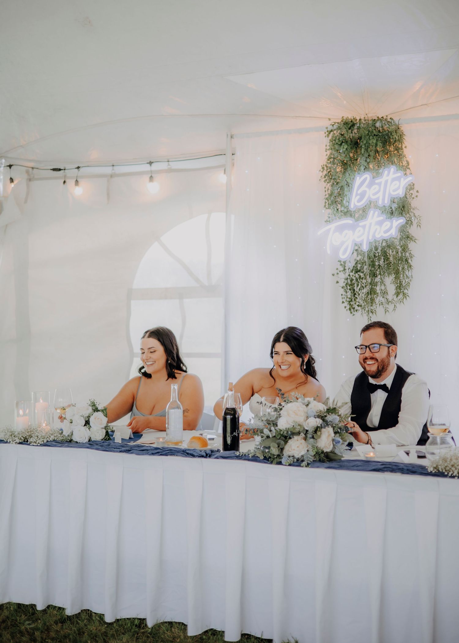 Wedding reception head table setup with white linens and string lights in a tented venue.
