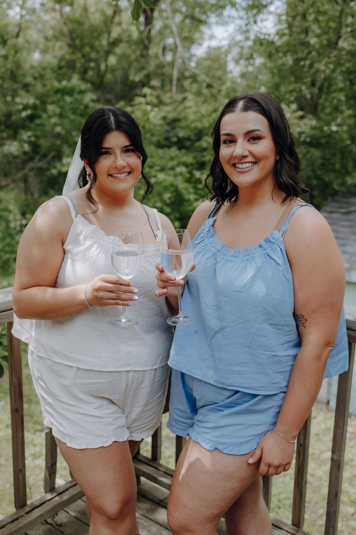 Two friends in casual summer outfits share drinks and laughter while relaxing on a wooden deck surrounded by greenery.