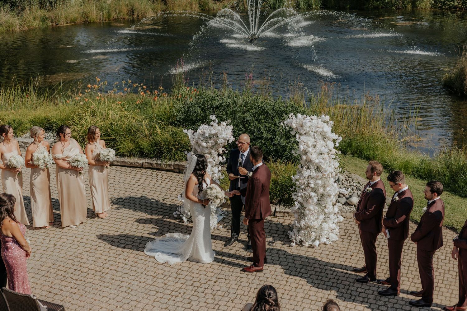 Wedding ceremony by a fountain with bridal party in burgundy and white attire.