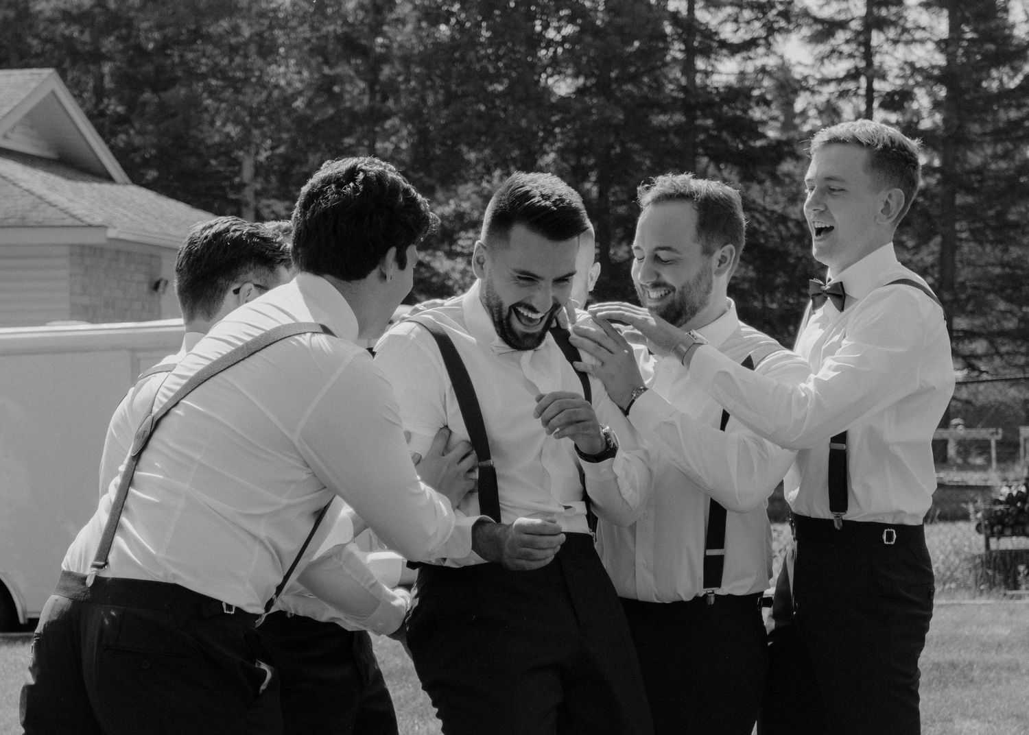 Groomsmen in suspenders and white shirts share a candid laughing moment together at an outdoor wedding.