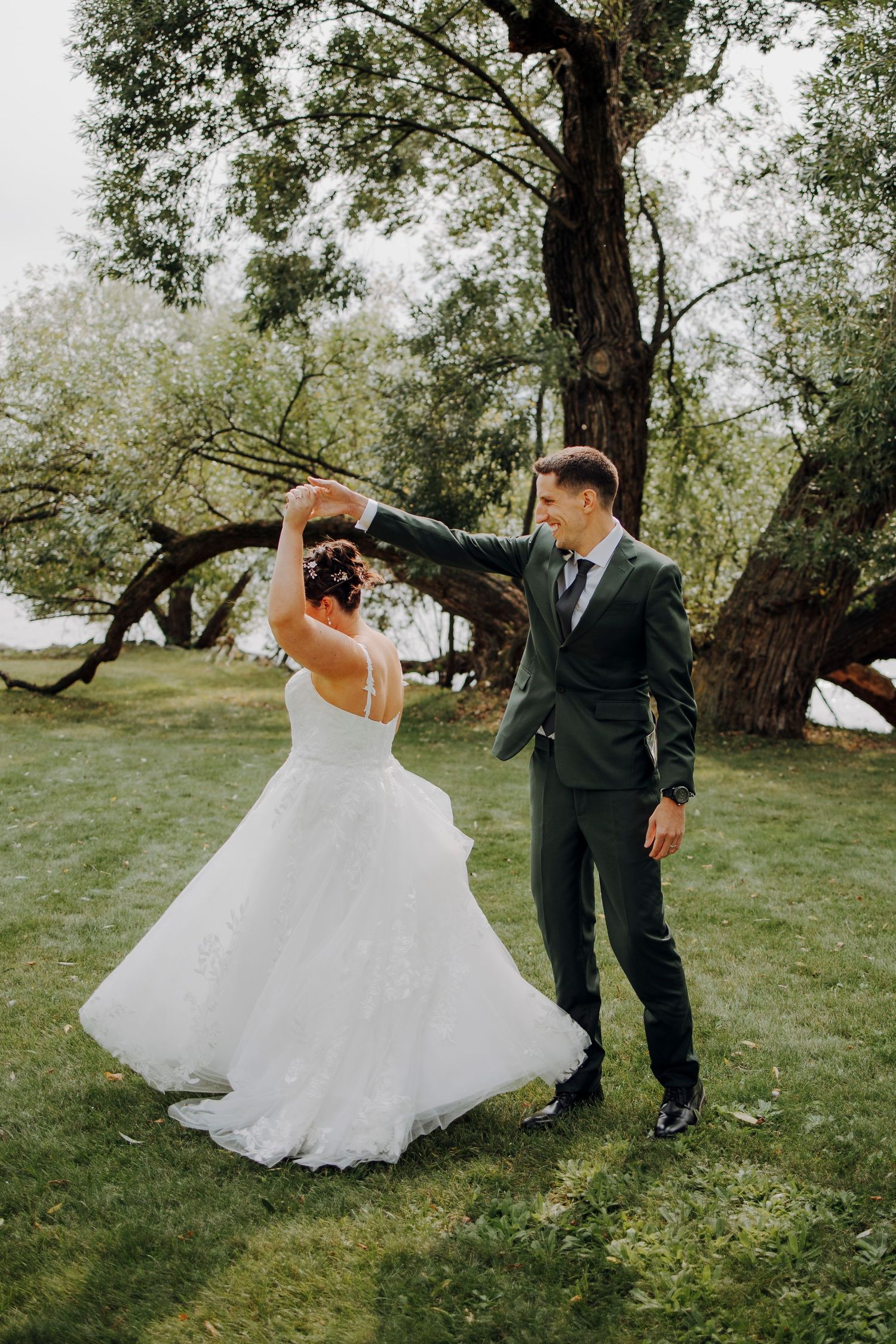 A couple in wedding attire dances and twirls playfully on green grass under trees.