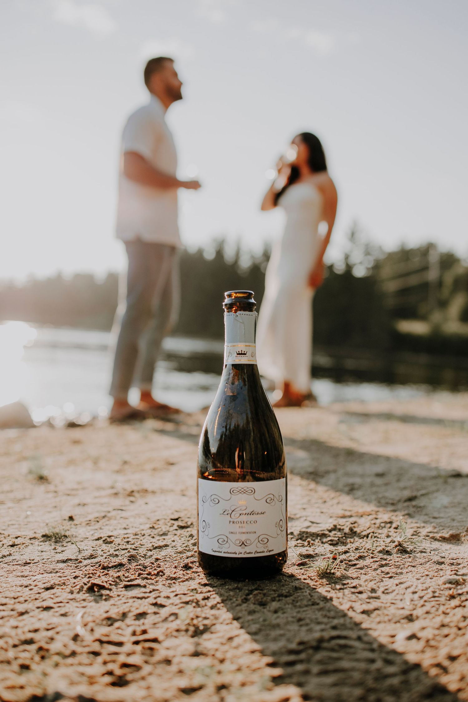 A bottle of champagne sits on beach sand with figures silhouetted in the background.