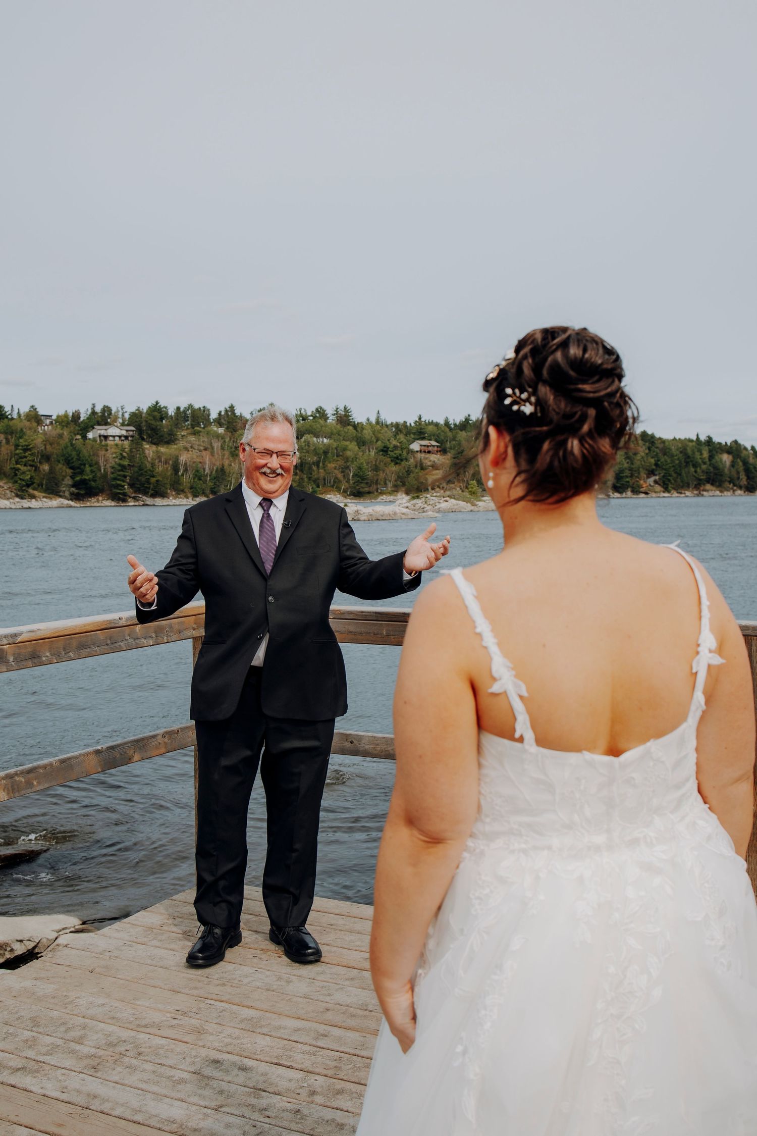 A wedding first look moment on a wooden dock by a lake with trees in the background.