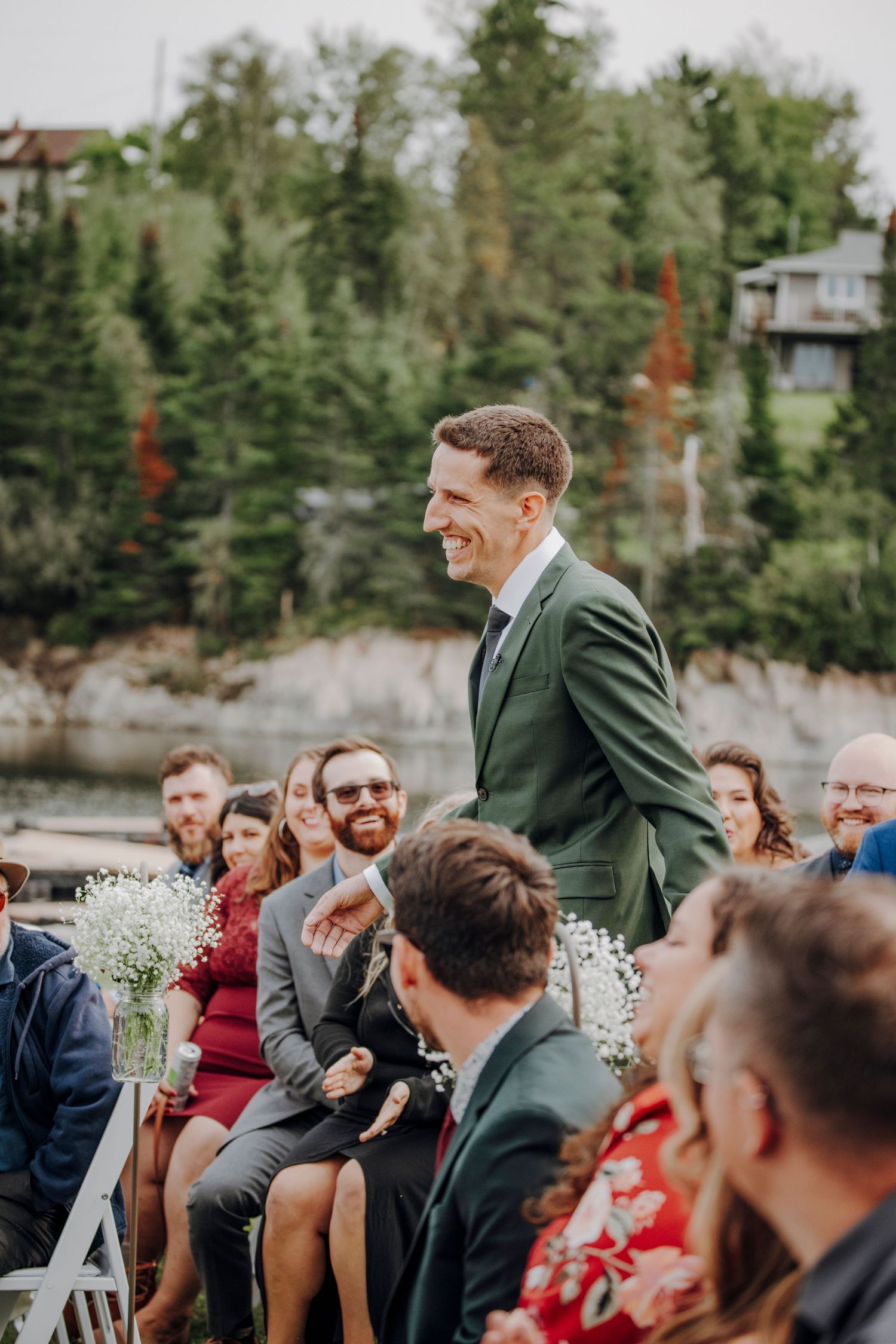 Wedding guests laugh and smile during an outdoor ceremony with white floral decorations and pine trees in the background.