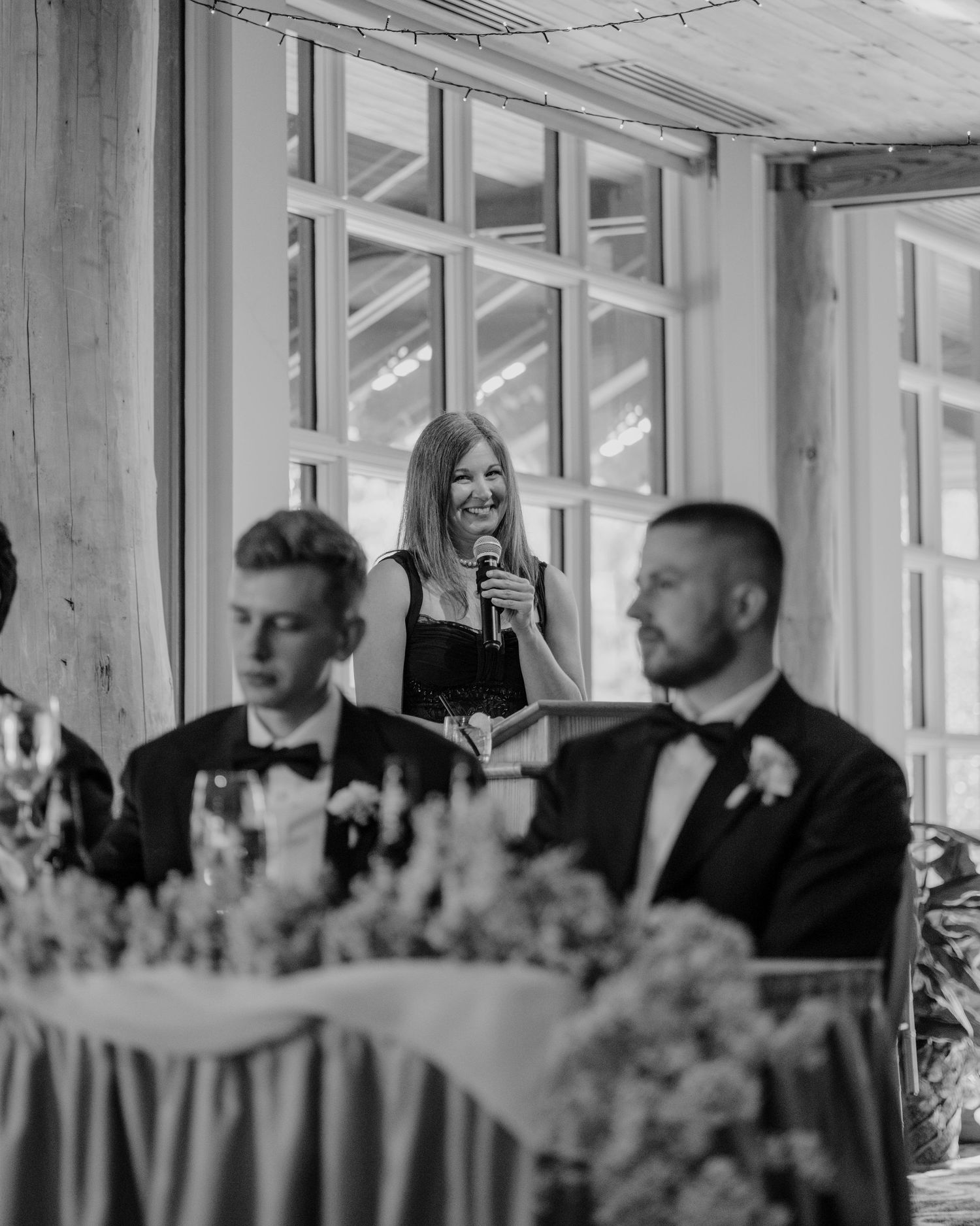 Black and white photo of wedding reception tables with floral centerpieces and guests in formal attire.