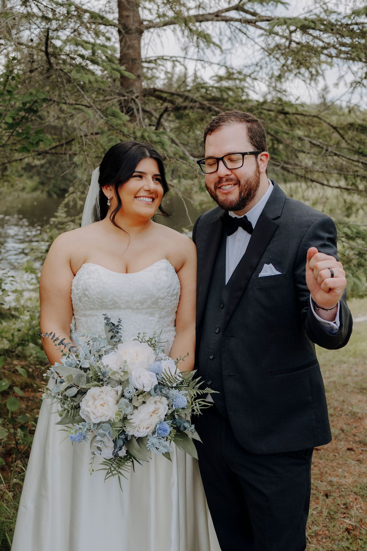Happy newlyweds pose together outdoors, with bride holding a pastel bouquet and groom in classic black tuxedo.
