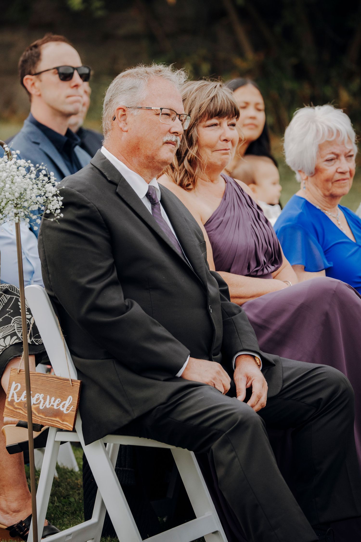 Wedding guests seated in white chairs during outdoor ceremony.