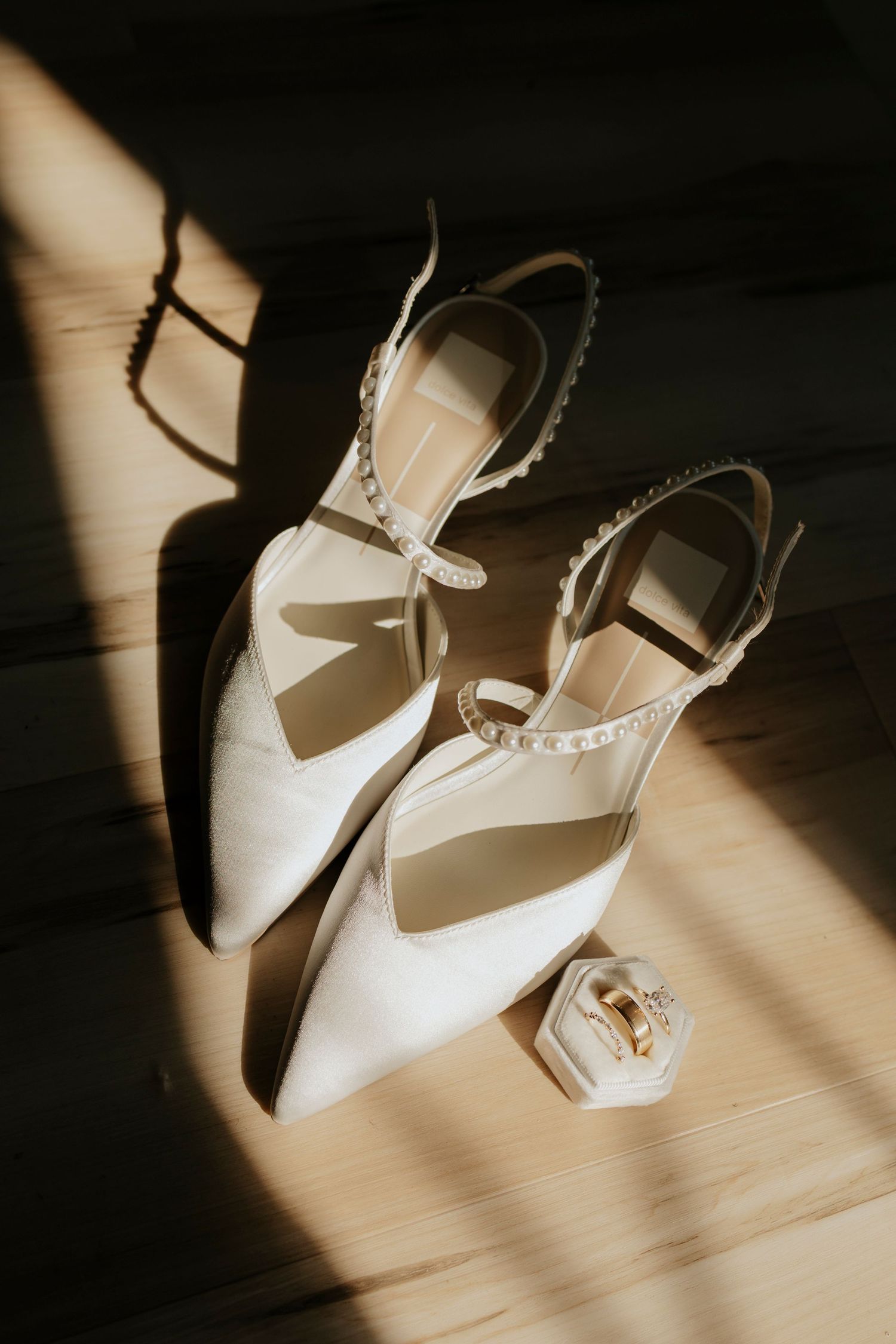 White bridal shoes and ring box casting dramatic shadows on a wooden surface in natural light.