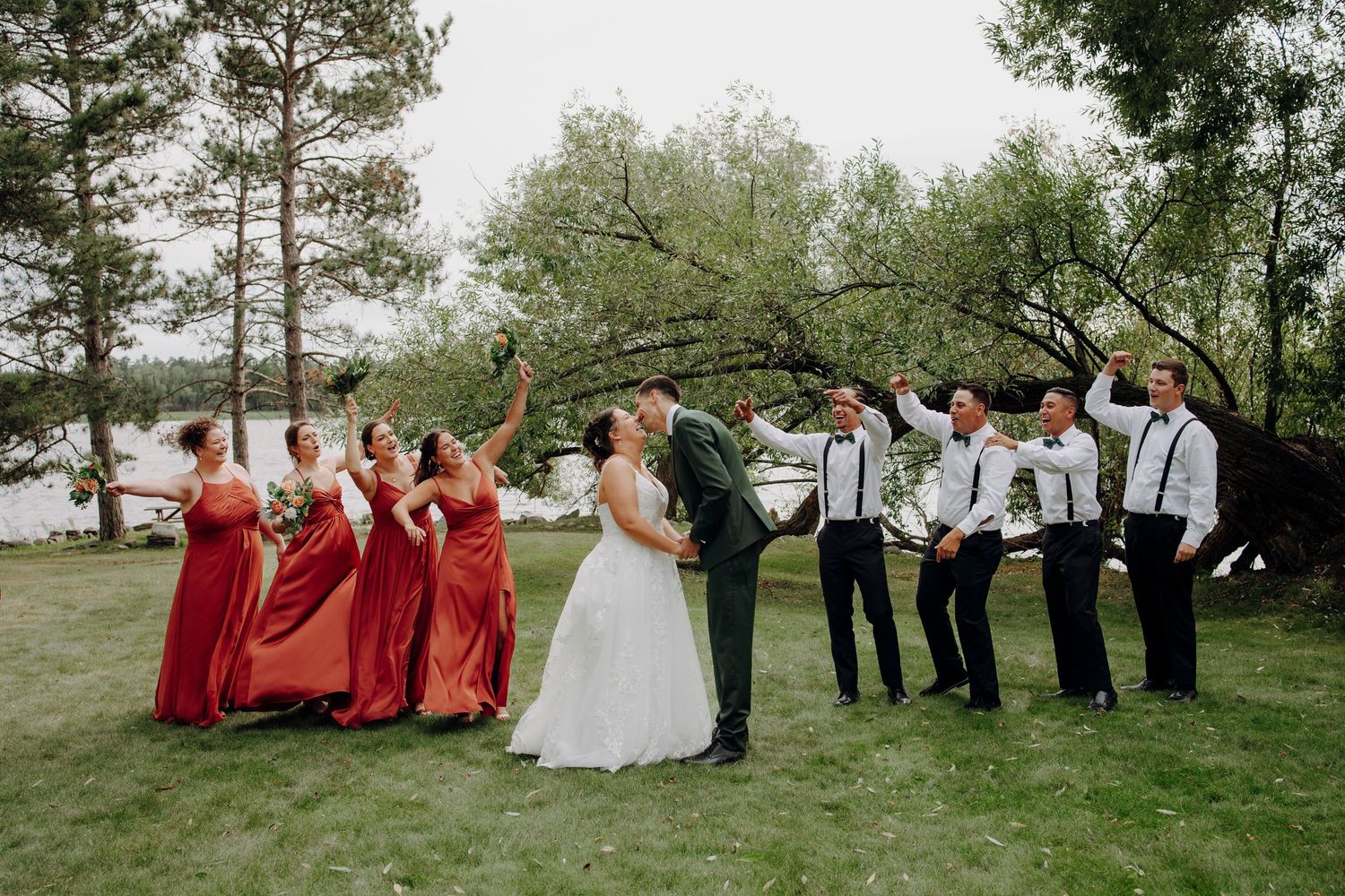 A panoramic shot of a wedding party gathered on a grassy hill with trees in the background.