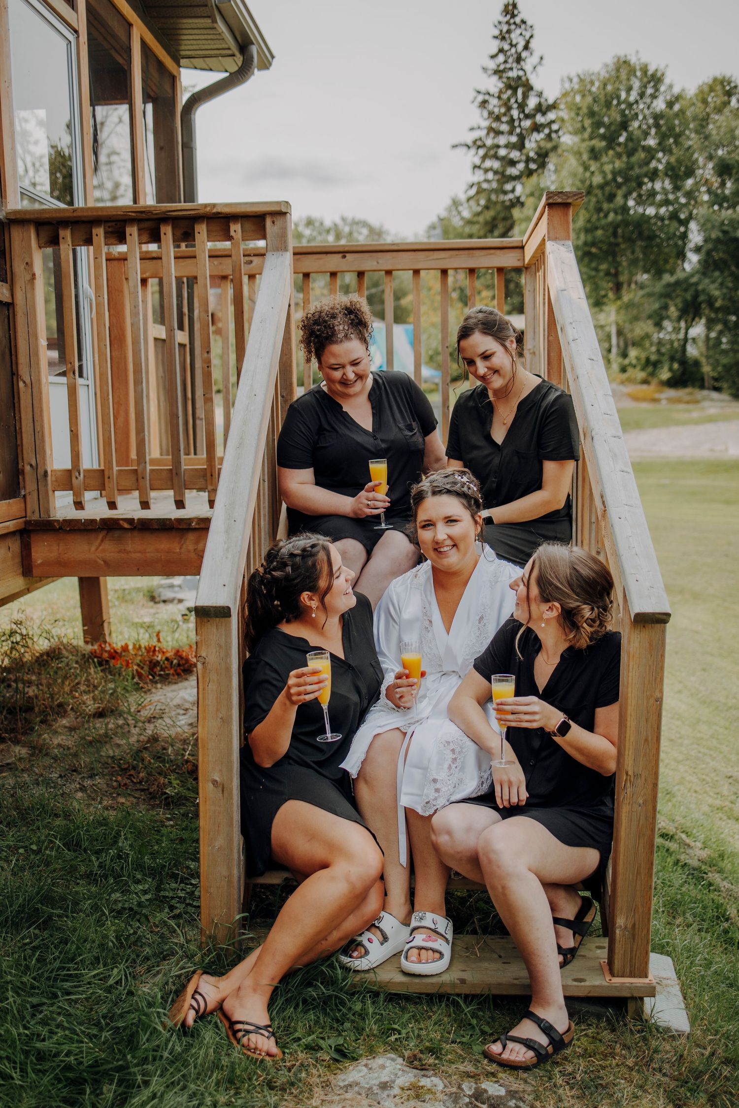 Group enjoying drinks and laughter together on wooden deck steps during outdoor gathering.