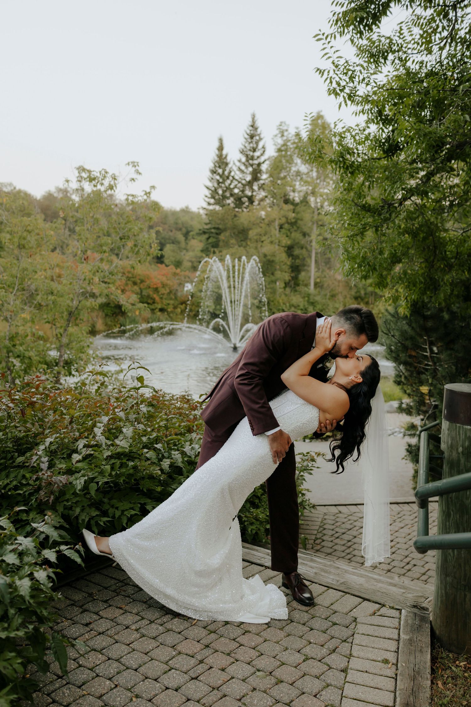 A couple in wedding attire performs romantic dips during their photoshoot by a lake.