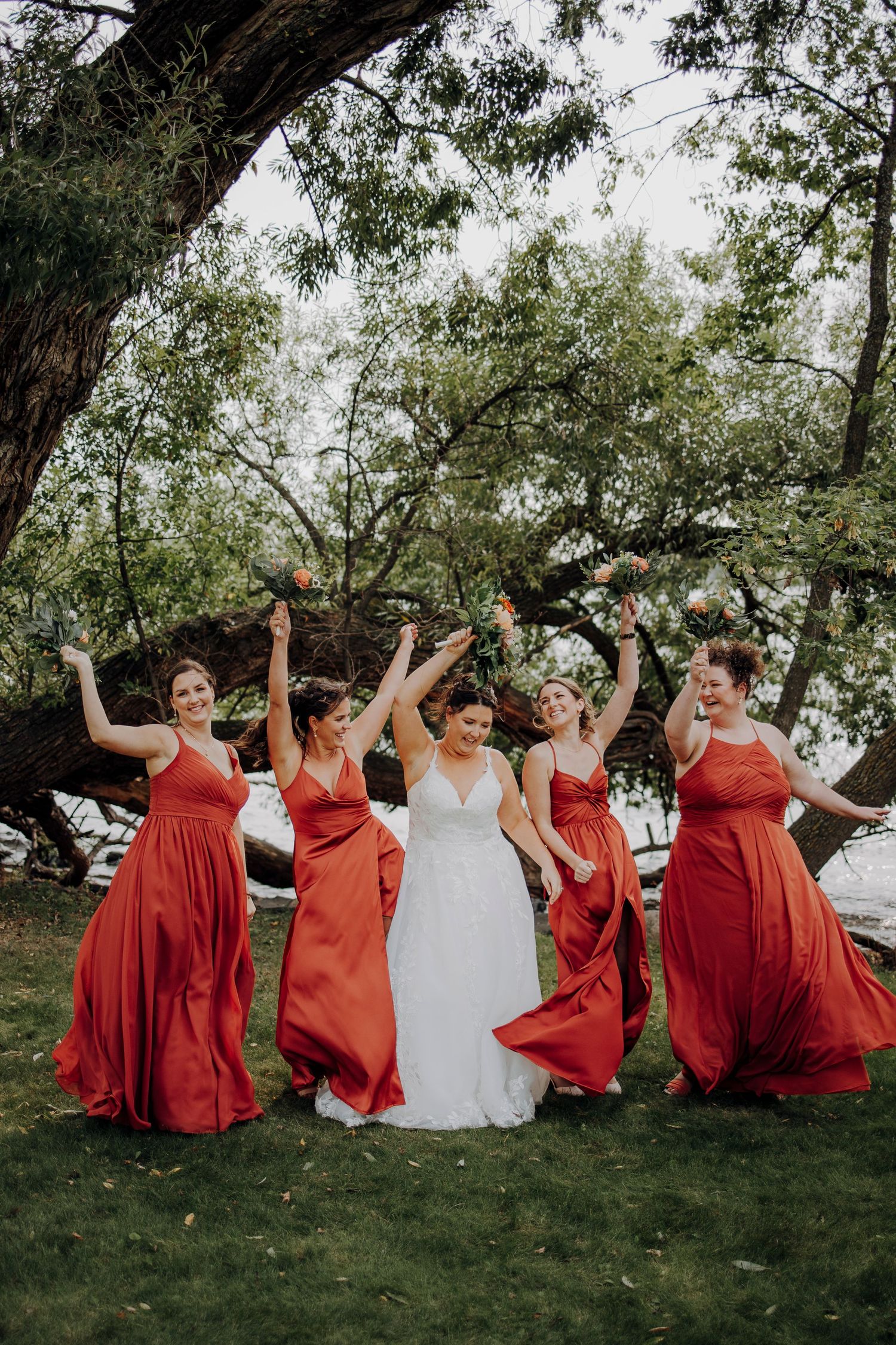 Bridesmaids in vibrant red dresses pose playfully on grass with the bride in white.