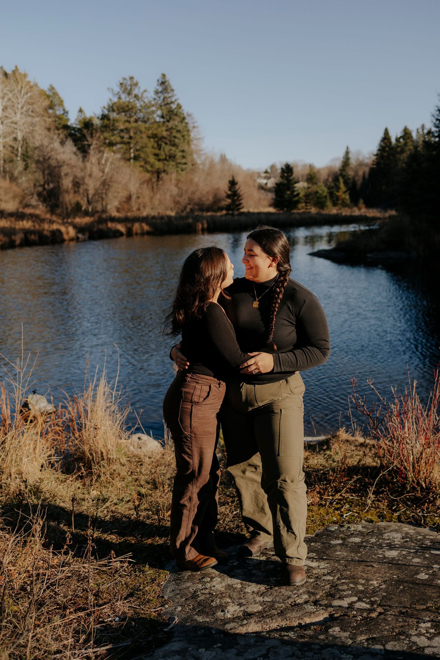 A couple shares a romantic moment by a serene lake surrounded by evergreen trees on a sunny afternoon.