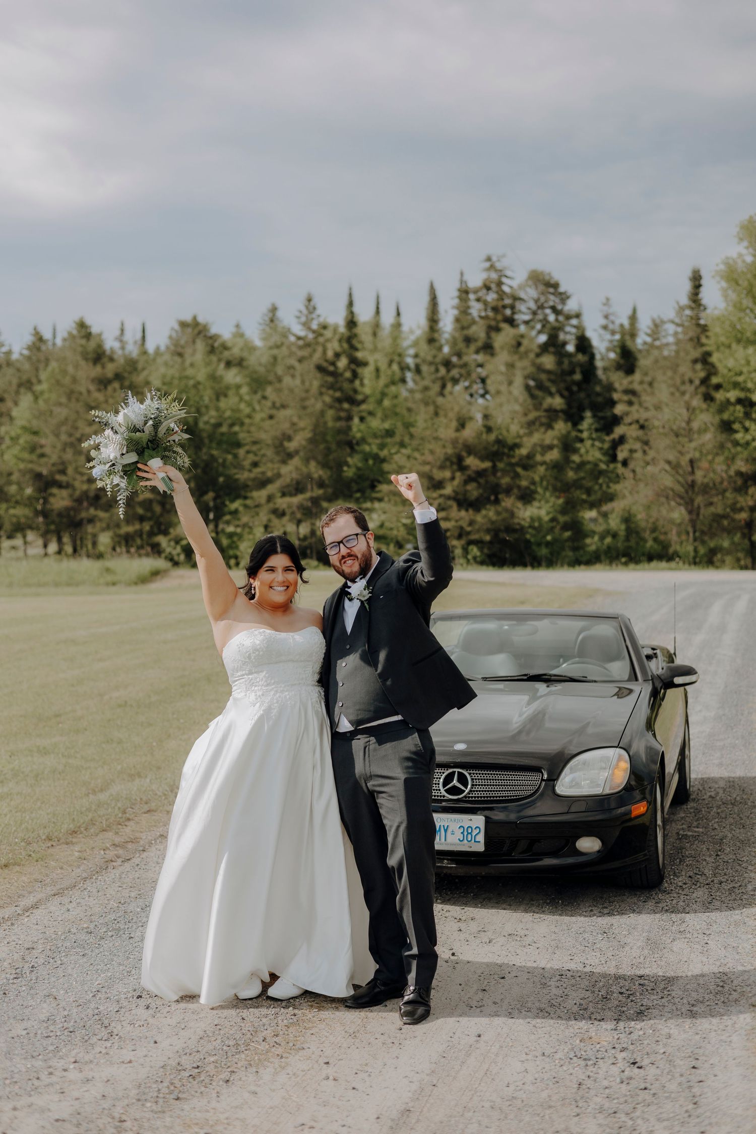 Wedding couple poses playfully next to a black luxury car on a country road.
