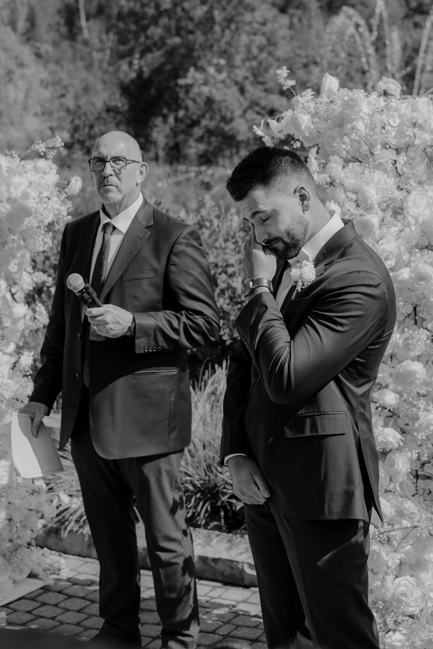 Two men in suits share an emotional moment during an outdoor wedding ceremony.