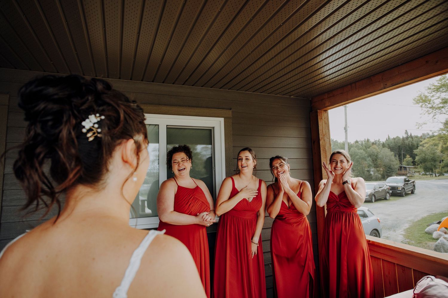 Bridesmaids in matching red floor-length dresses react with excitement on a covered porch.