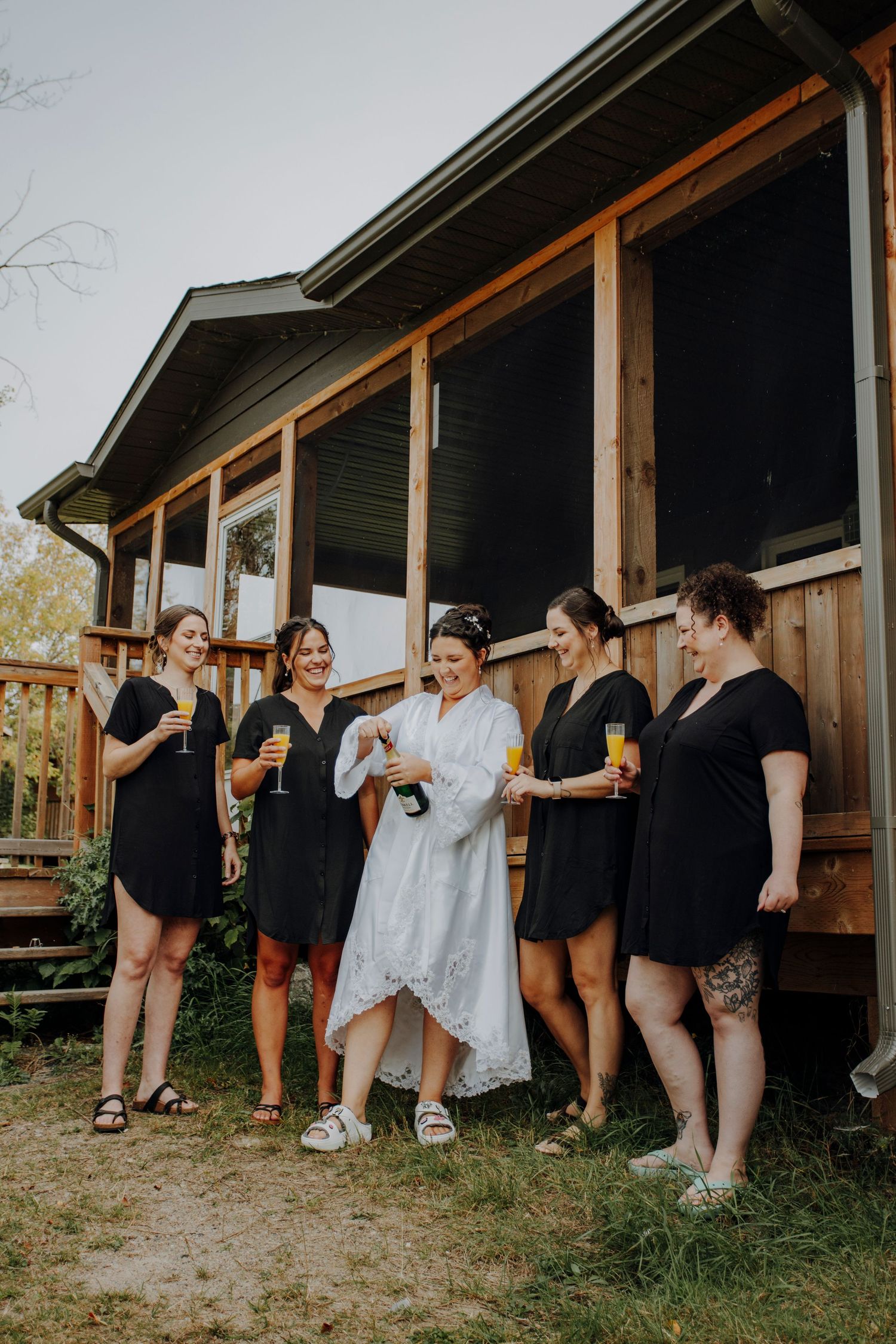 Wedding party poses playfully behind rustic wooden cabin in coordinated black and white outfits.