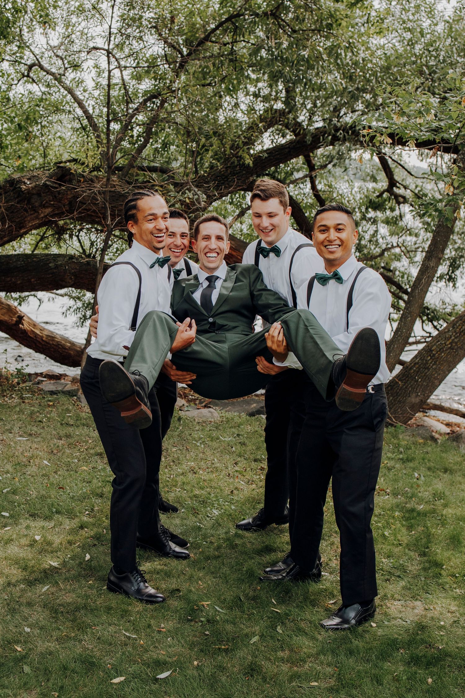 Groomsmen in suspenders and bowties playfully holding up the groom in an outdoor setting.