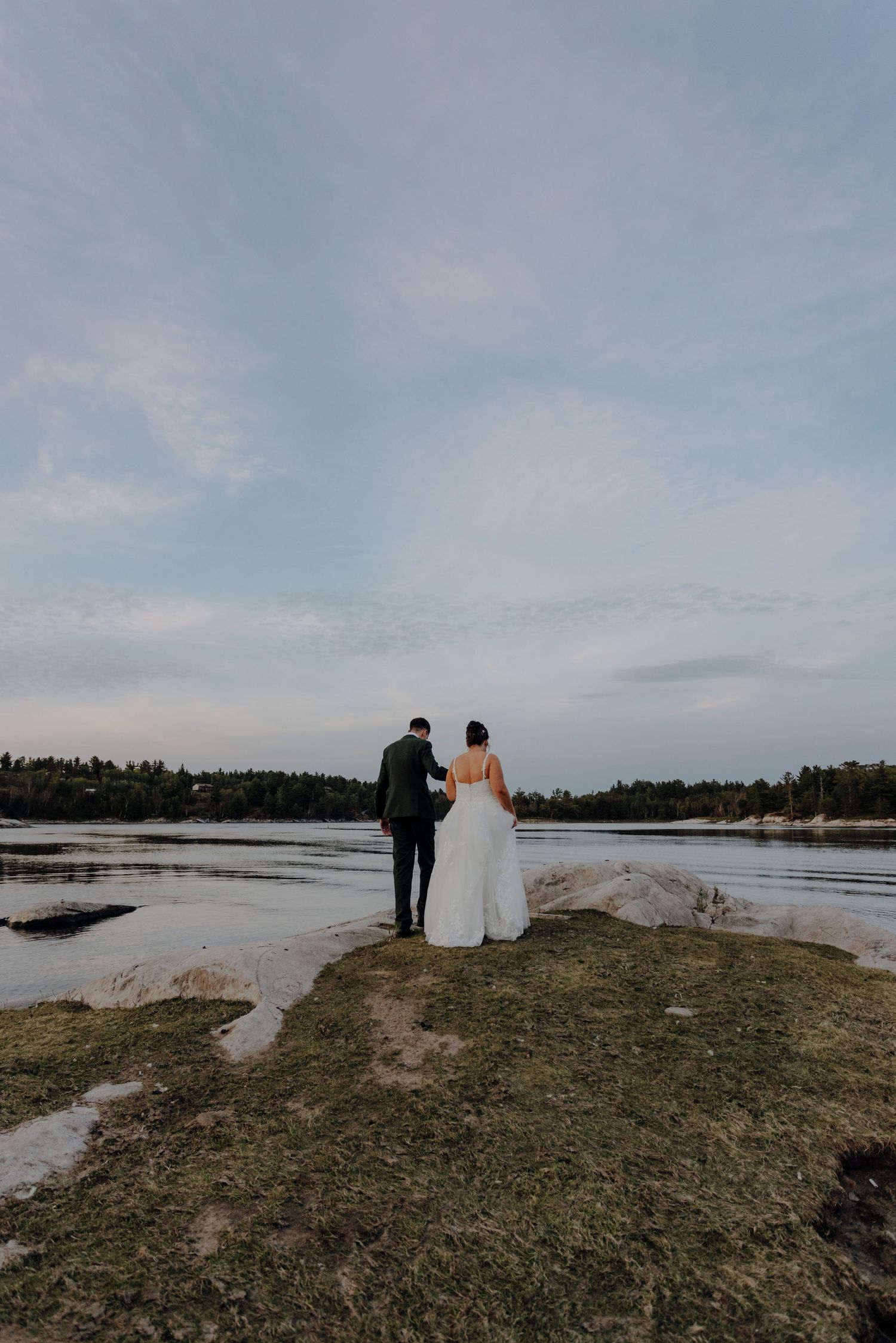 Silhouetted wedding couple stands on rocky shoreline against cloudy sky and water.