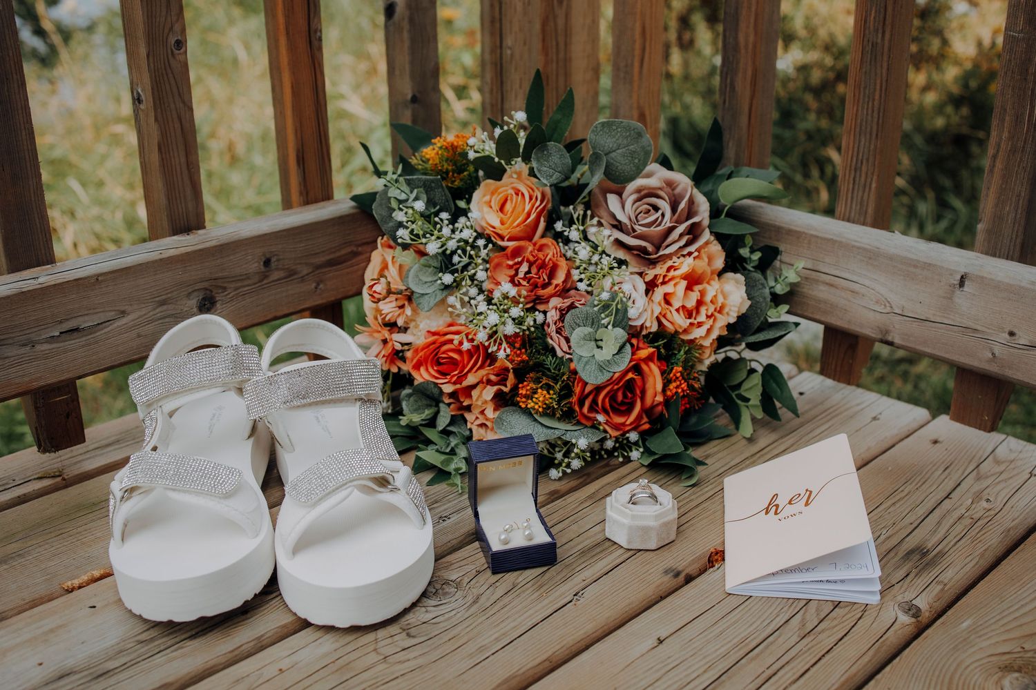 Wedding details arranged on rustic wooden deck with floral bouquet, white shoes, invitation and teacups in warm lighting.